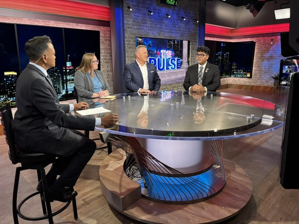 A television talk show set with four people seated around a glass table, two men and two women, with a city skyline backdrop and a screen displaying 'The Pulse.'