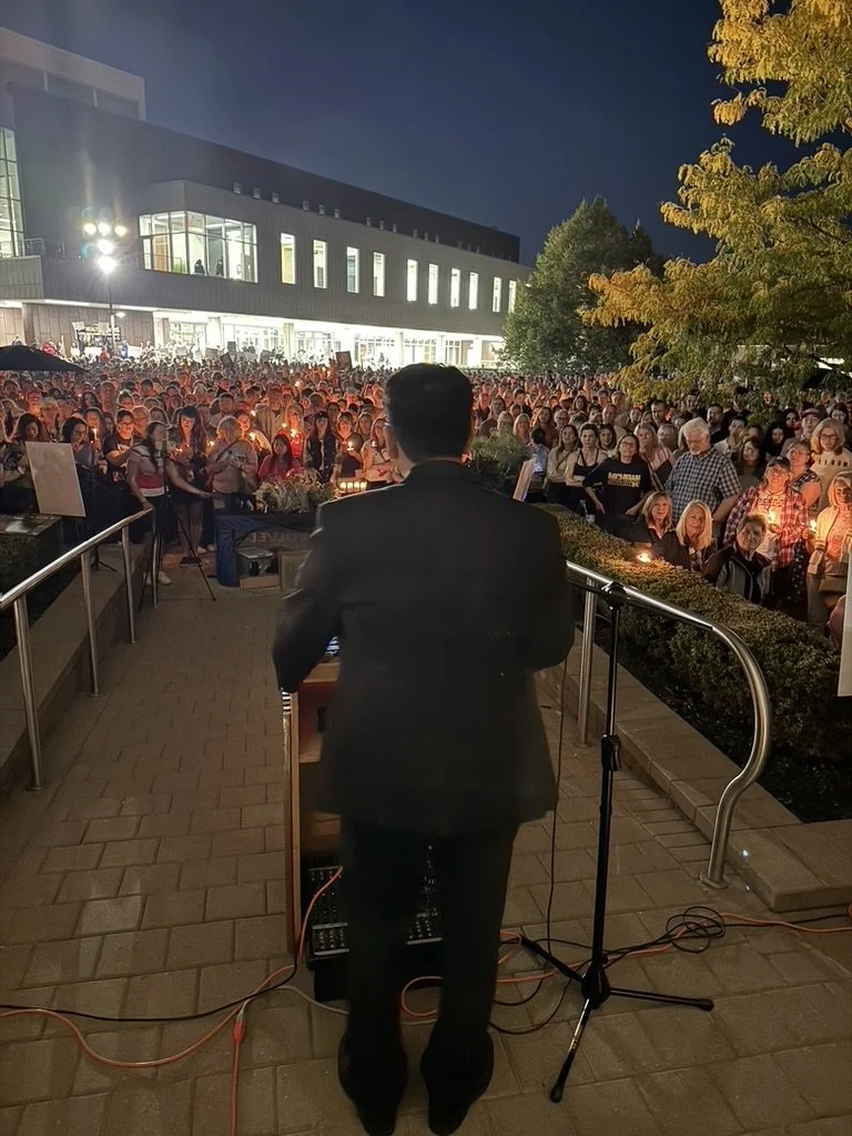 A man in a suit performs at a podium outdoors during the evening, facing a large crowd gathered for an event or vigil, with a building and trees illuminated in the background.