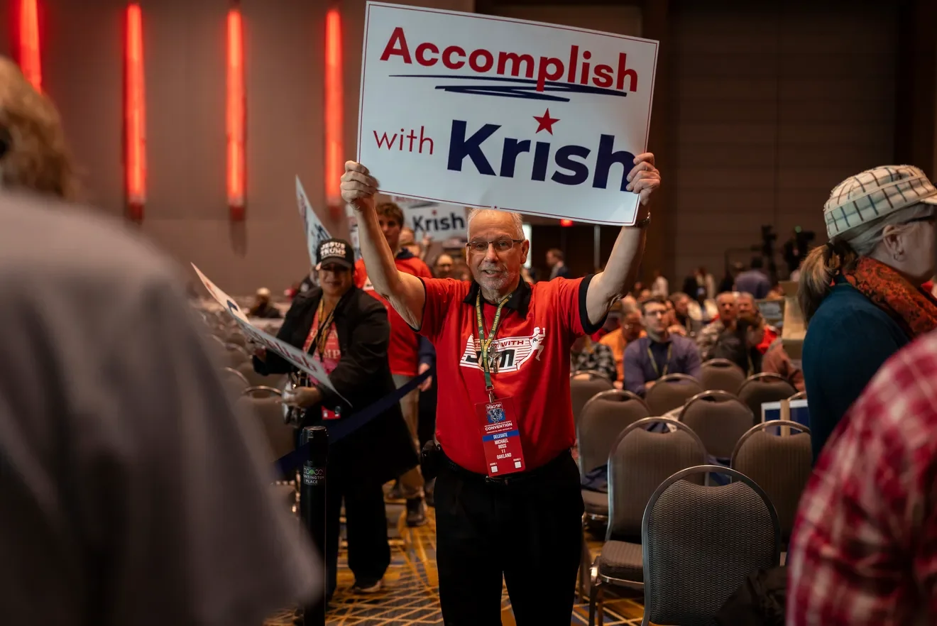 A supporter holding a sign that says 'Accomplish with Krish' at Michigan GOP Convention with many seated attendees.