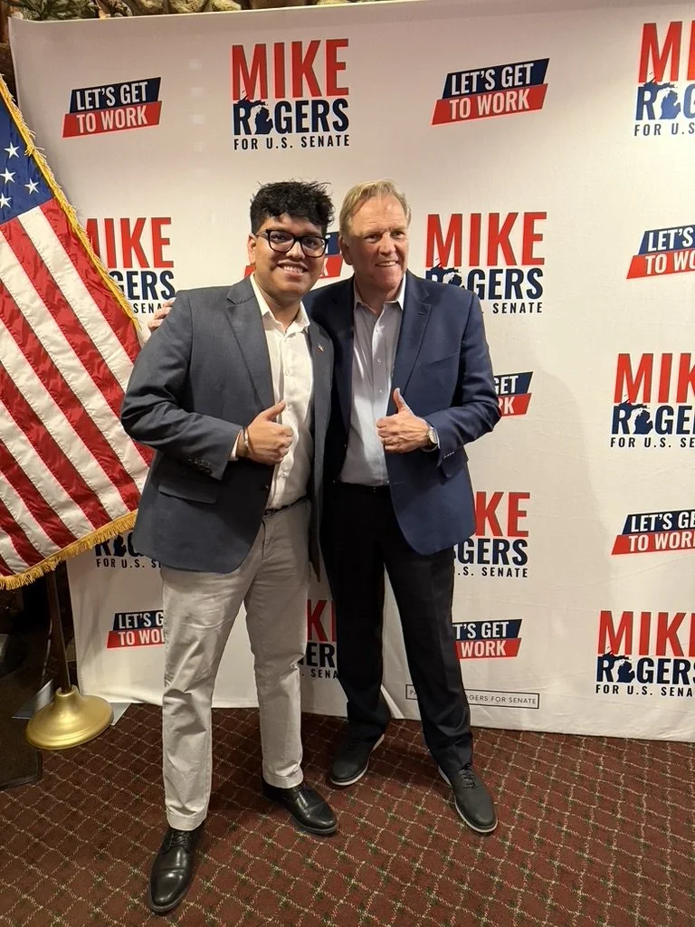 Two men in suits standing in front of a campaign backdrop with text promoting Mike Rogers for U.S. Senate, and an American flag on the left. Both men are smiling and giving thumbs-up.