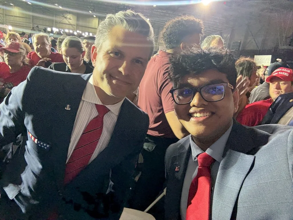 Two men in suits and red ties taking a selfie at a crowded indoor event, with many people in the background wearing red.