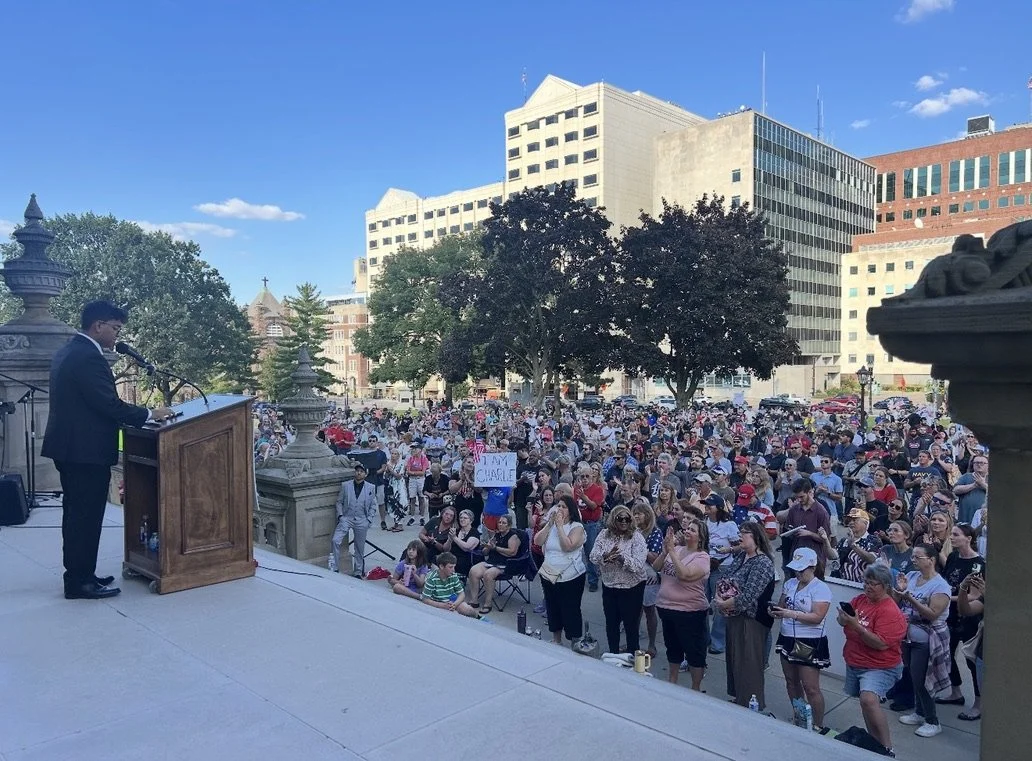 A man in a suit speaking at a podium on an outdoor stage in front of a crowd gathered in a city square with tall buildings and trees.