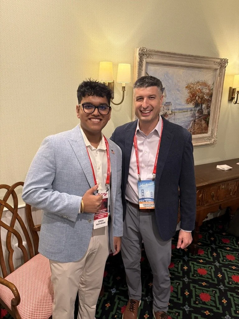 Two men standing in a room, smiling at the camera. One man is wearing glasses and a light-colored blazer, the other is in a dark blazer. They have event badges around their necks. Behind them is a classical painting of a landscape with water and tree