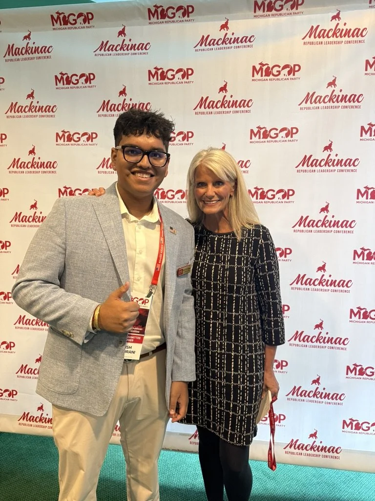 Young man and woman standing together at the Mackinac Republican Leadership Conference with a red and white backdrop featuring the conference name and logo. They are smiling, and the man is giving a thumbs-up.