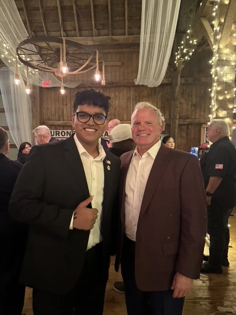 Two men in suits smiling at a gathering in a rustic wooden barn decorated with string lights and drapes.