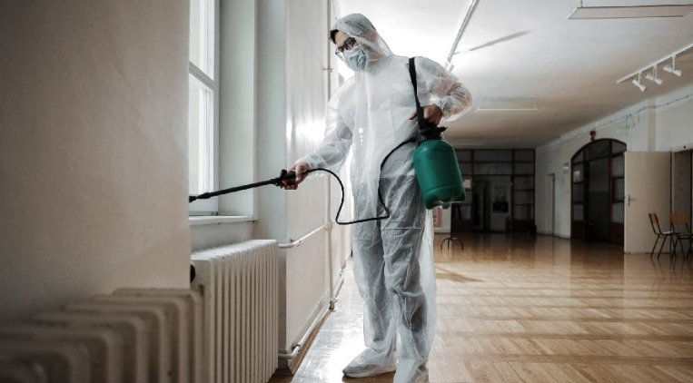 Person in protective suit disinfecting a windowsill with a sprayer in a spacious room with wooden floors.