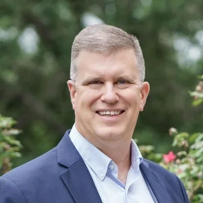 A smiling man in a blue suit and light blue shirt standing outdoors with green trees and flowers in the background.