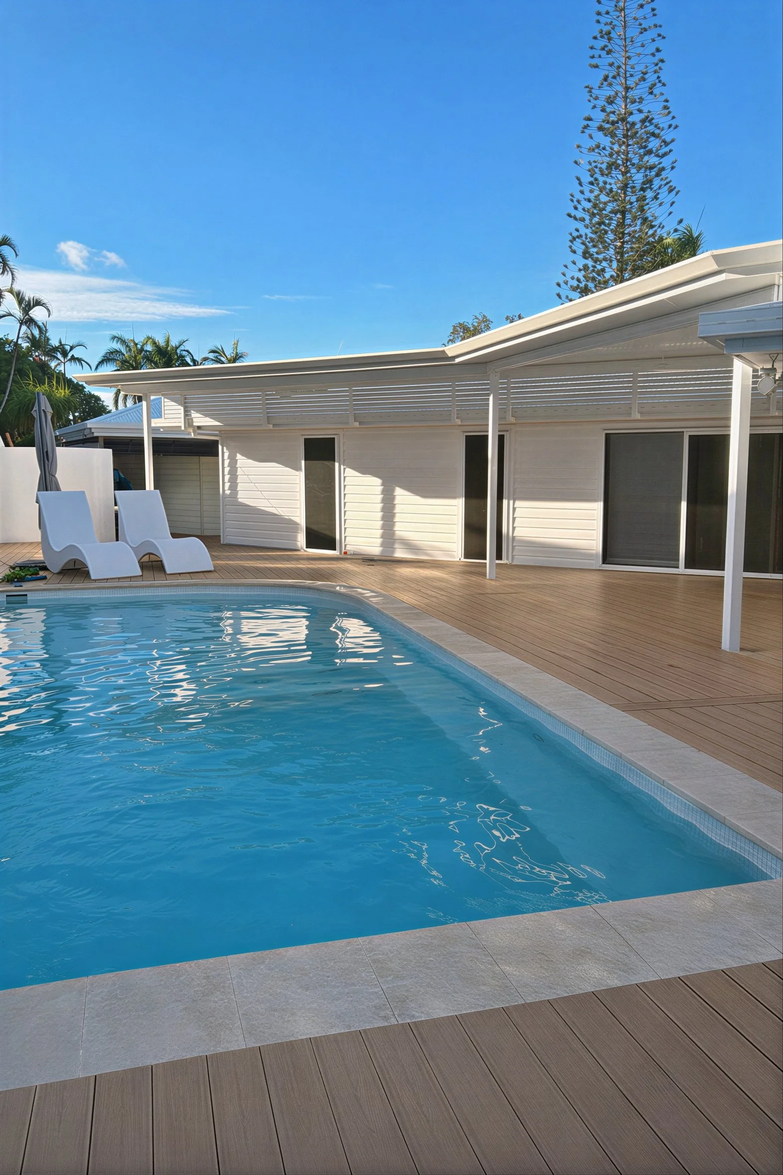 A backyard with a swimming pool, two white lounge chairs, a closed umbrella, and a deck with wooden flooring. There's a white house with large sliding glass doors and a slanted roof, and trees in the background under a clear blue sky.