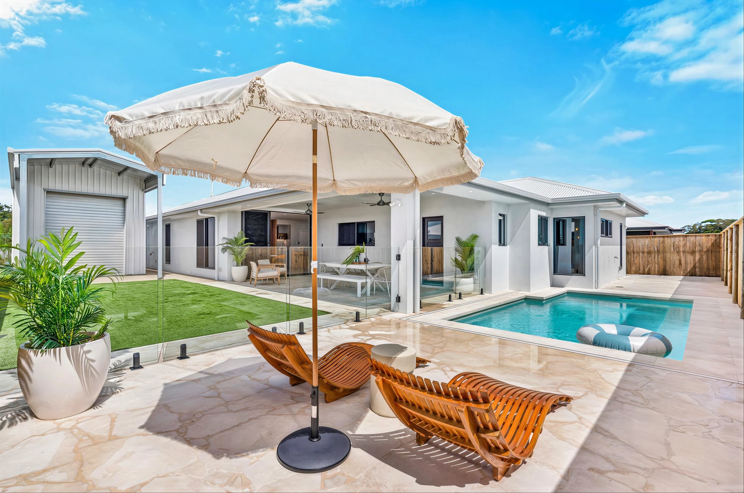 Custom concrete pool featured in a modern backyard, complemented by two wooden lounge chairs beneath a large white umbrella, potted plants, and a sleek glass fence. The setting includes a white home, manicured lawn, and a separate garage building.