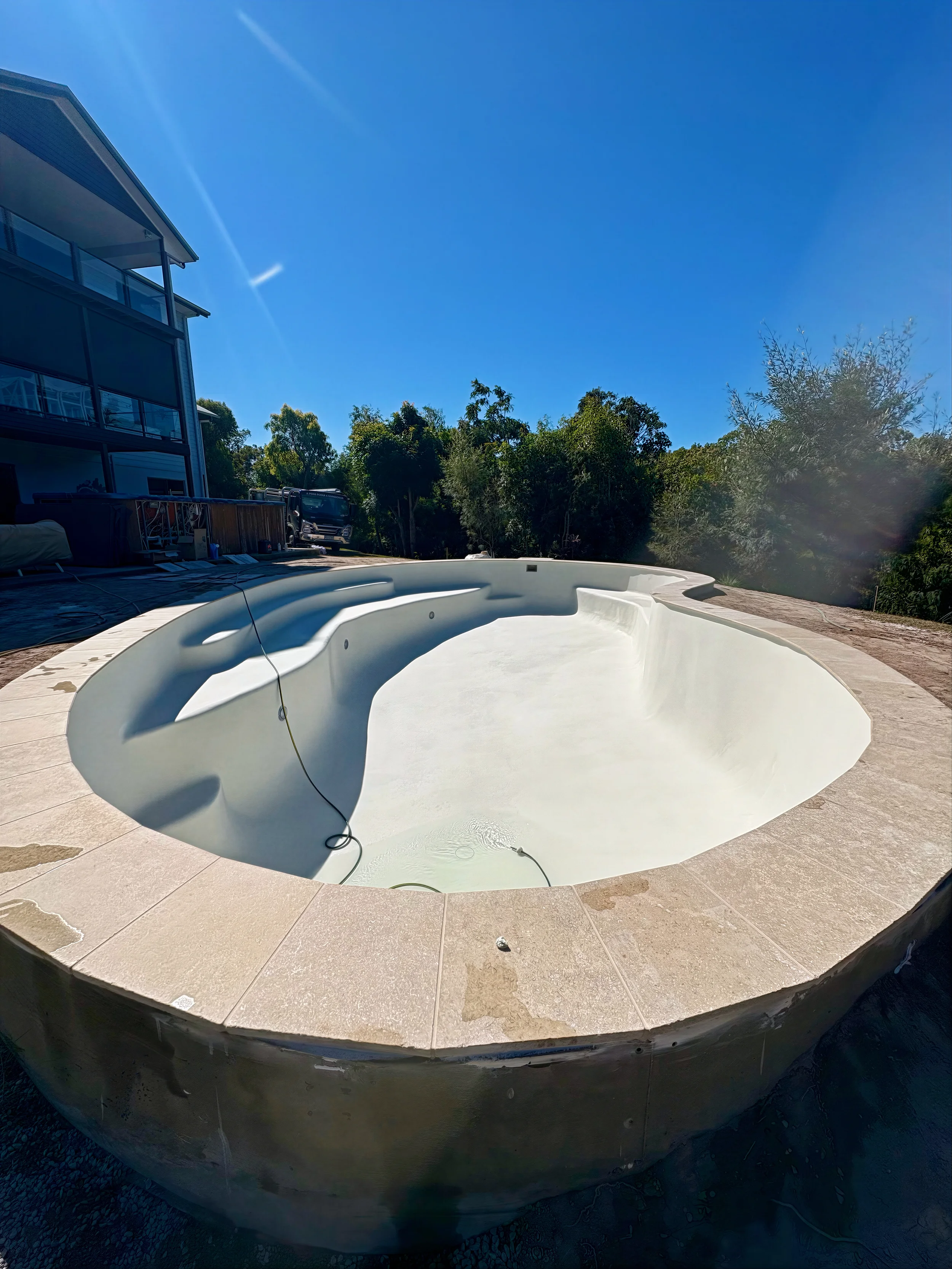 Custom concrete pool with a distinctive curved design and tiled border, shown empty in a sunny backyard under a clear blue sky, with a house to the left and trees in the background.