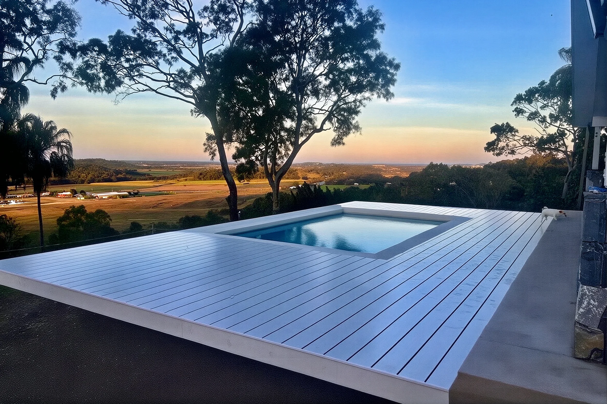 Custom concrete pool set beside a spacious modern deck on a hillside, overlooking rural properties and surrounded by lush trees.