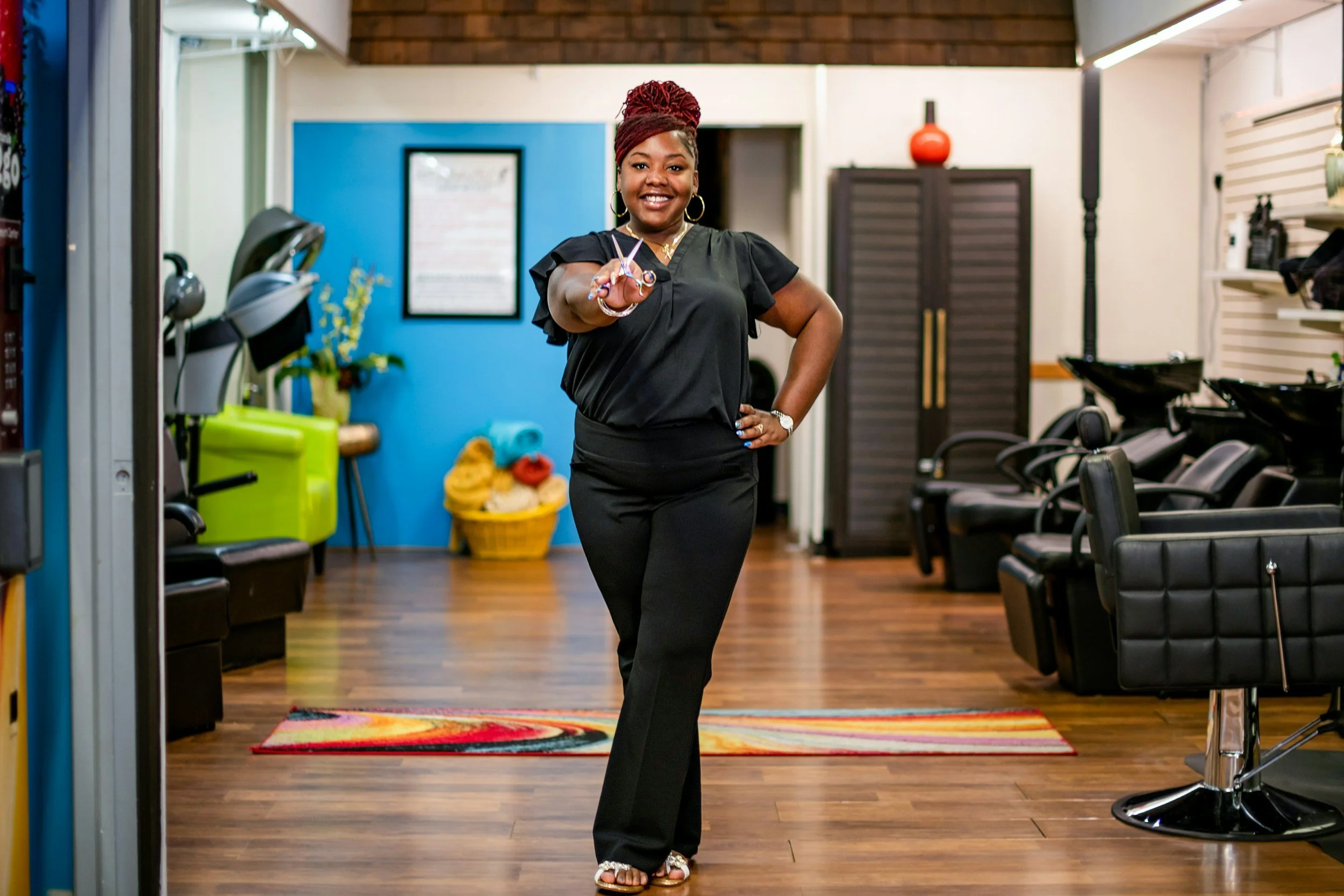 Woman in black outfit standing inside a hair salon, smiling, pointing towards the camera, with hairdressing chairs and equipment in the background.