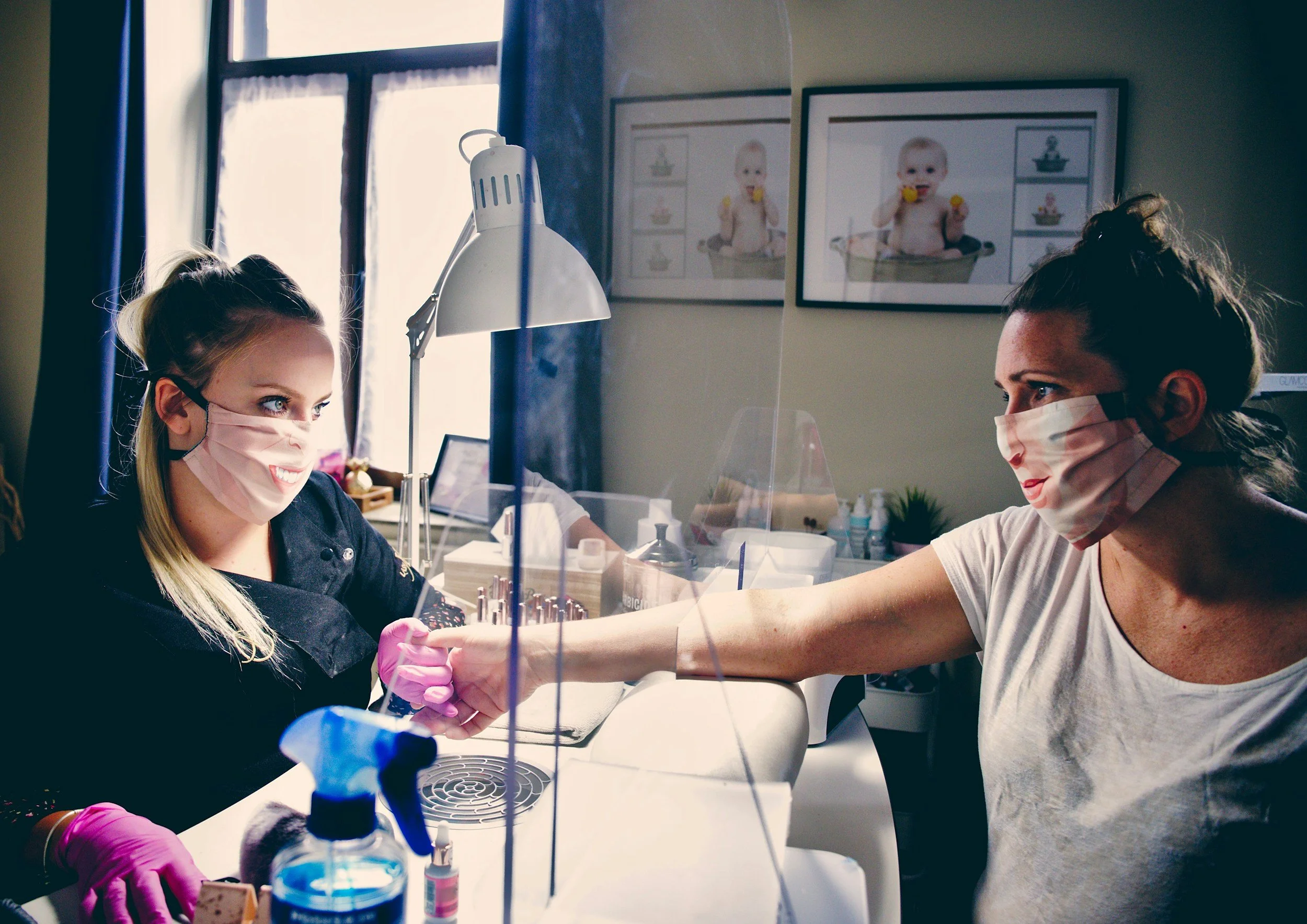 A woman in a white shirt and face mask receives a manicure from a woman in a black top and face mask, wearing pink gloves, in a nail salon with framed pictures of babies on the wall.