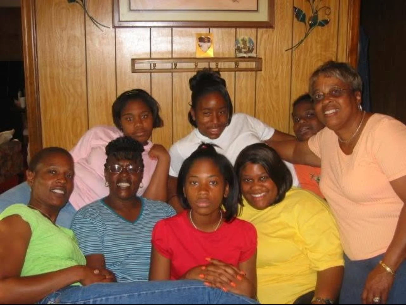 Group of nine women and girls gathered together indoors, smiling and posing for the camera, with wood-paneled walls in the background.