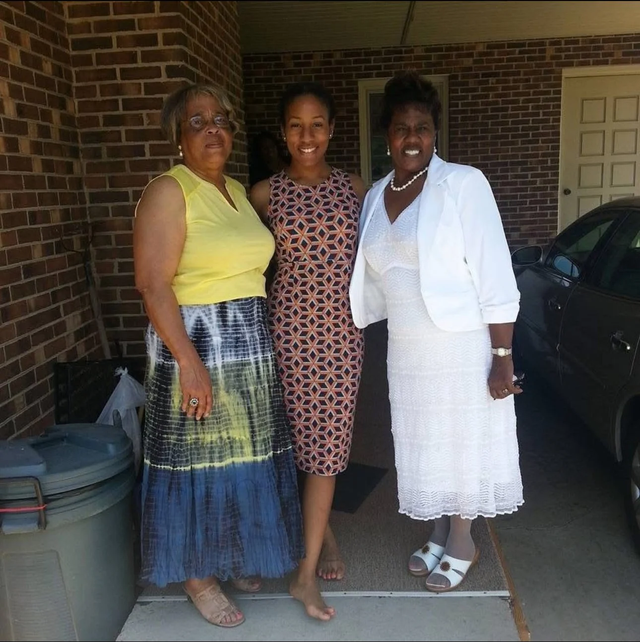 Three women standing outside a brick house, posing for a photo. The woman on the left wears a sleeveless yellow top and a long patterned skirt. The woman in the middle wears a sleeveless geometric patterned dress. The woman on the right wears a white dress and jacket with white sandals.