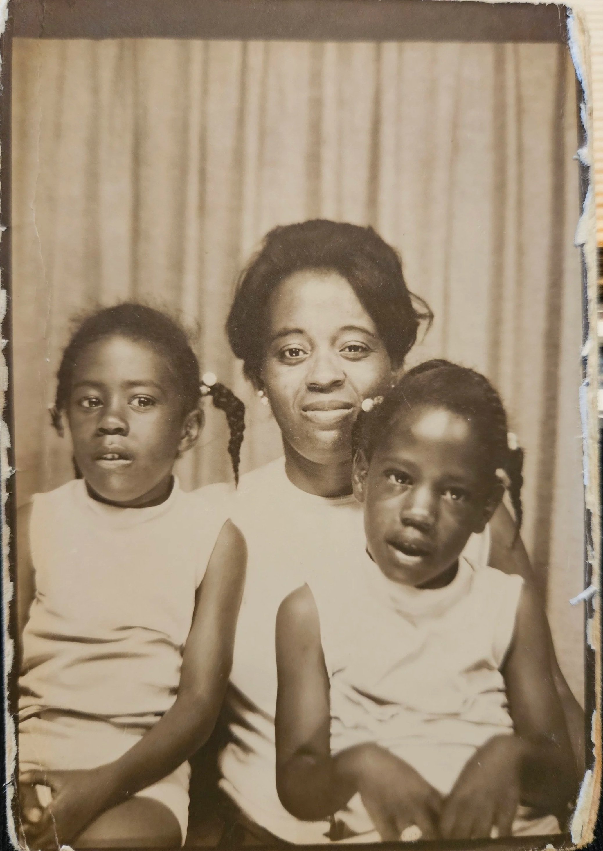Black-and-white photograph of a woman with two young girls, all wearing sleeveless tops, sitting in front of a curtain background.