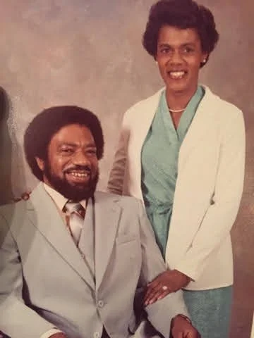 A man and woman in formal clothing posing for a photo, with the man sitting and the woman standing beside him, both smiling.