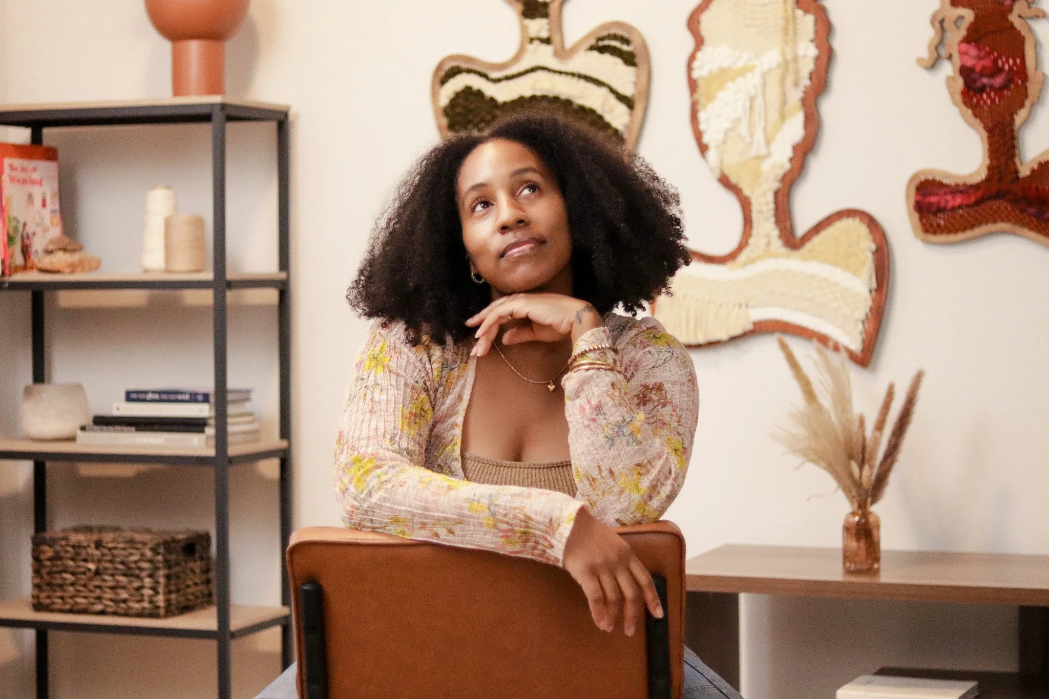 A young woman with curly hair sitting at a wooden table, resting her chin on her hand, looking upwards. The background features textured art pieces on the wall and a shelving unit with books and decorative items.