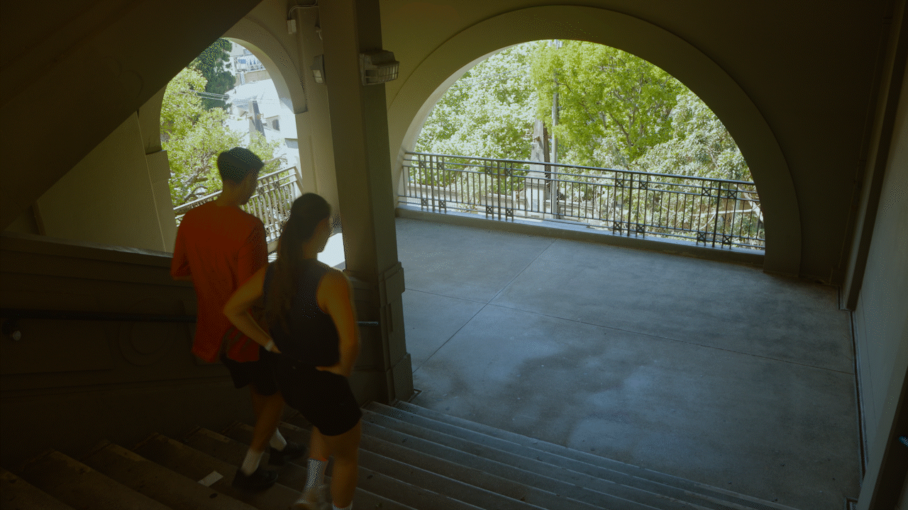 A man and a woman walking down concrete stairs in an outdoor stairwell, with a view of trees and buildings seen through arched openings.