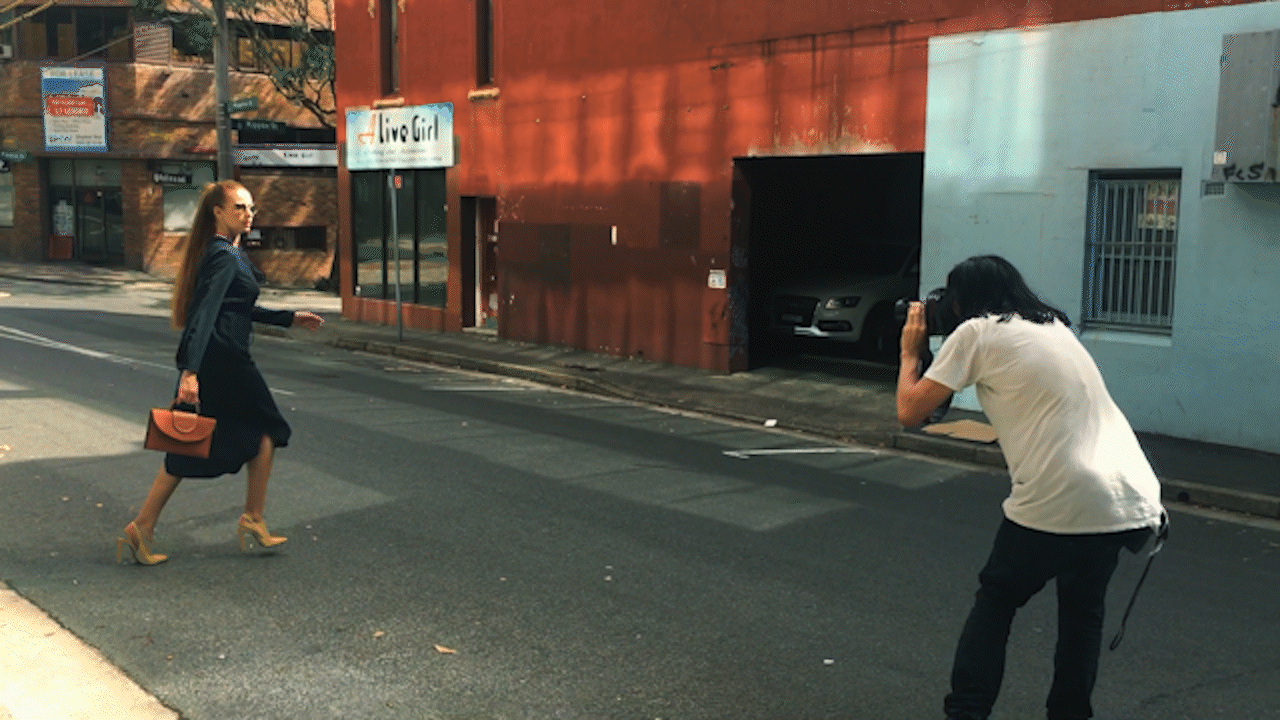 A woman in a black dress and high heels walking across the street while a photographer takes her photo. The woman carries a small handbag. The background shows a red building with signs and a garage.