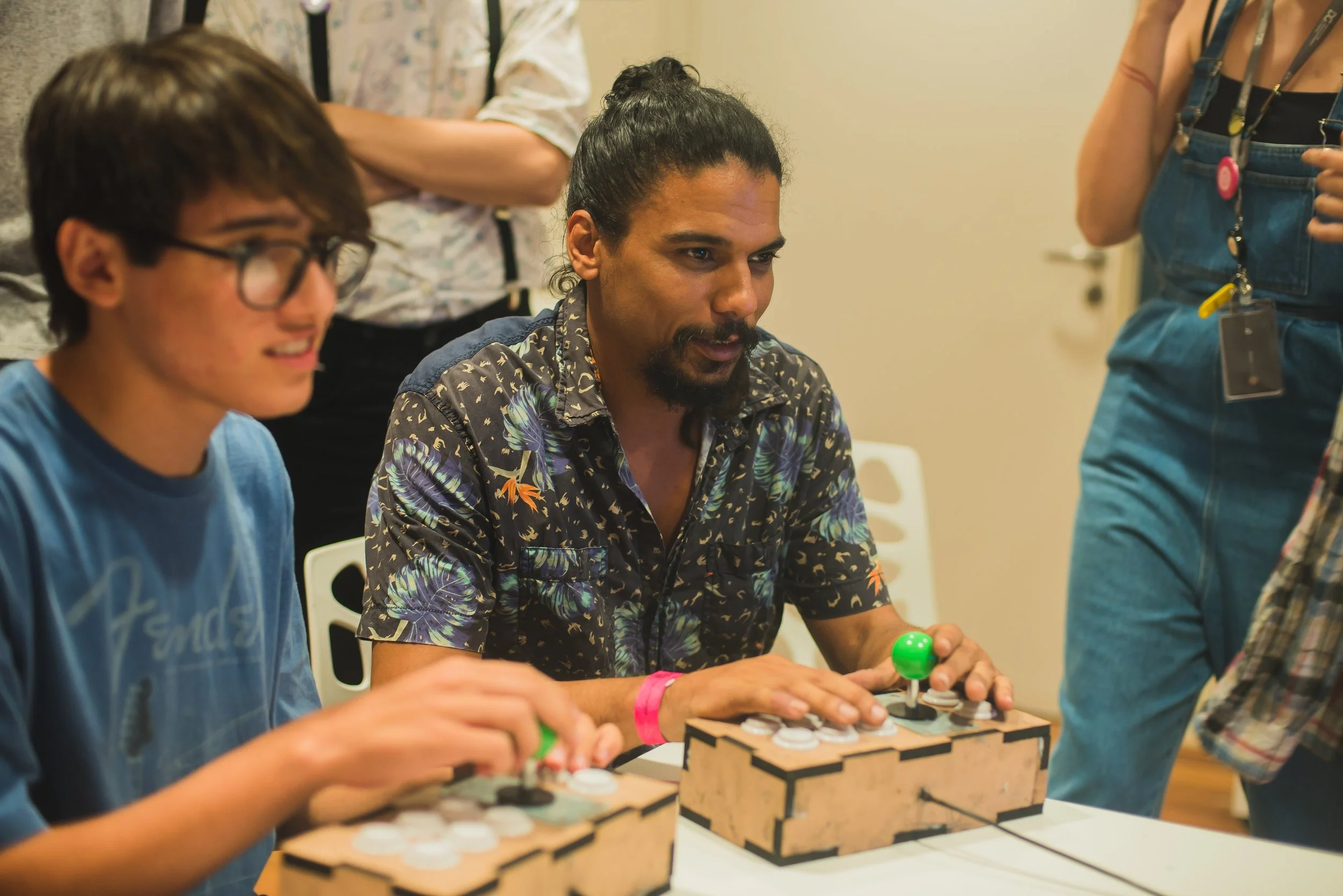 Homem e jovem jogando videogame de arcade com botões e joystick, em uma mesa, em ambiente interno.