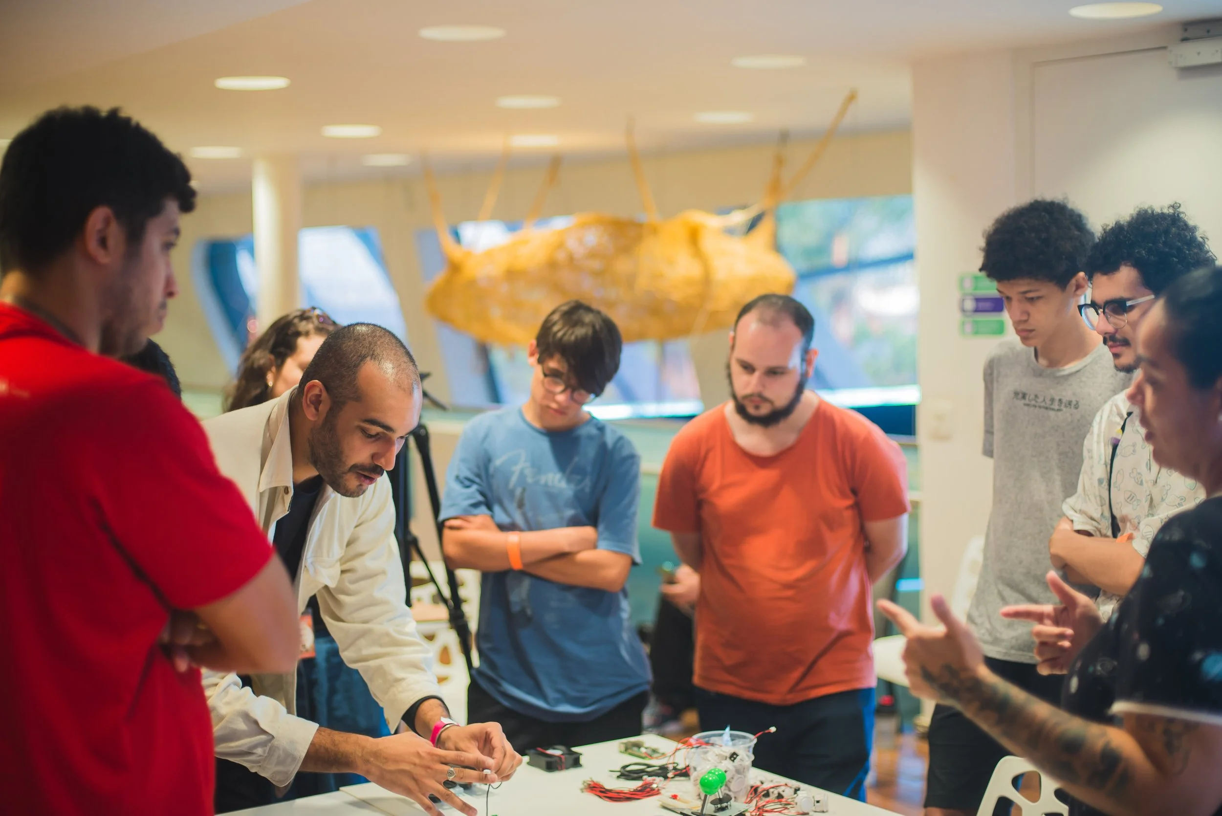 Grupo de pessoas participando de uma oficina de eletrônica, manipulando componentes eletrônicos em uma mesa, com ar de concentração, em um ambiente bem iluminado