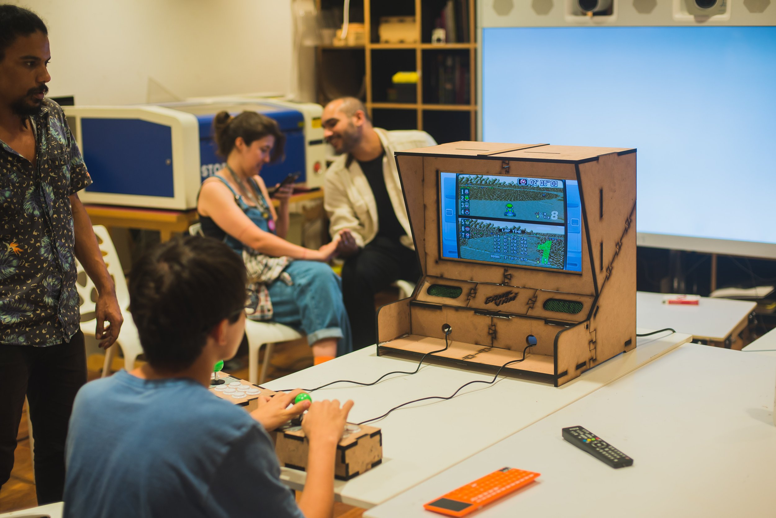 Pessoa jogando um videogame arcade feito de madeira em um evento, com uma tela em destaque e outras pessoas ao fundo em um ambiente interno.