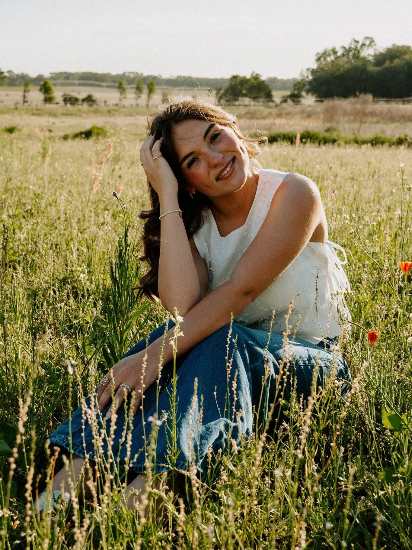 A young woman with brown hair and freckles sitting in a grassy field during daytime, smiling at the camera with her hand on her head, wearing a white sleeveless top and blue jeans, surrounded by wildflowers with a background of open landscape and trees.