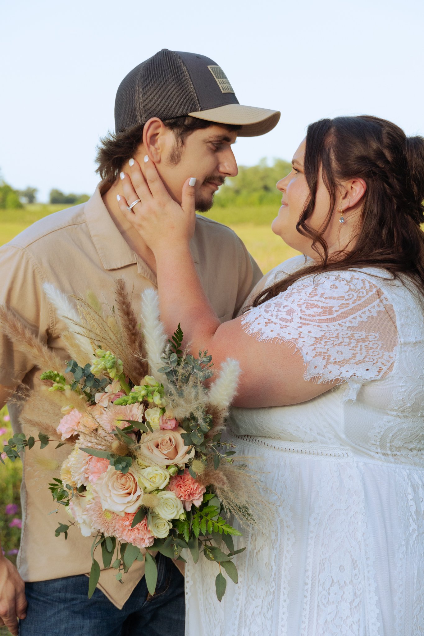 A couple in outdoor wedding attire sharing a moment, with the woman holding a large bouquet of pink, white, and green flowers.