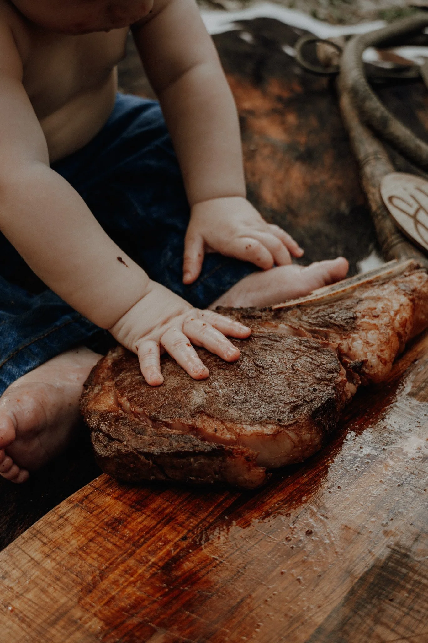A child with bare arms and wearing blue shorts is touching a large roasted beef with their hands, on a wooden cutting board.