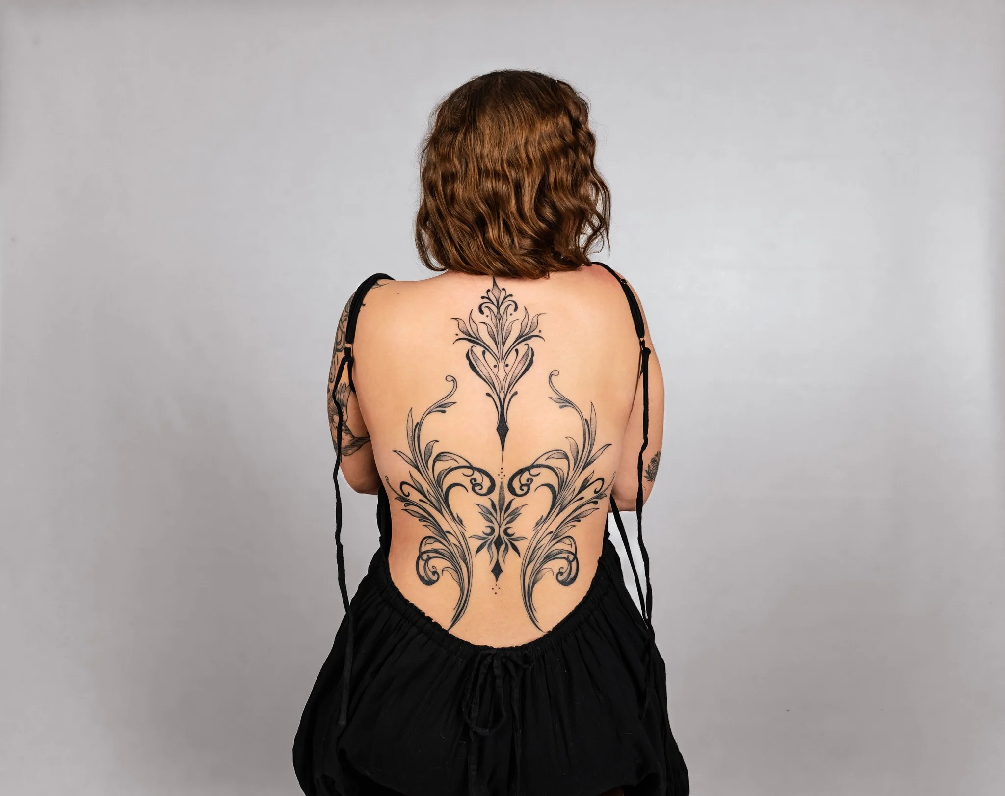 Back view of a woman with shoulder-length brown hair, wearing a black dress, showing a large floral tattoo on her back and tattoos on her arms, against a plain gray background.