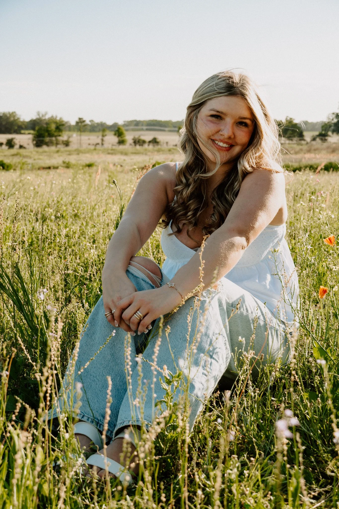 A young woman with long, wavy blonde hair sitting in a grassy field, smiling at the camera, wearing a white dress and white shoes, with a scenic background of trees and open land under a clear sky.