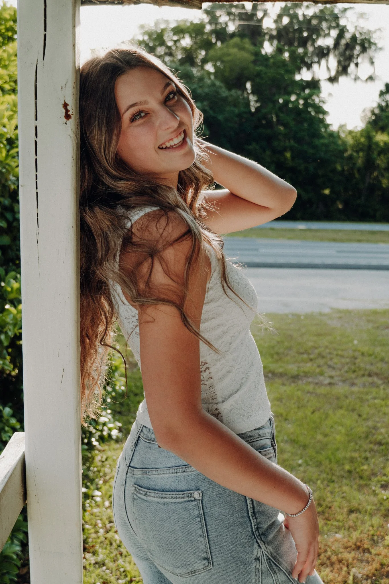 A young woman with long wavy brown hair smiling and leaning against a white wooden post outdoors during daytime, with trees and a grassy area in the background.
