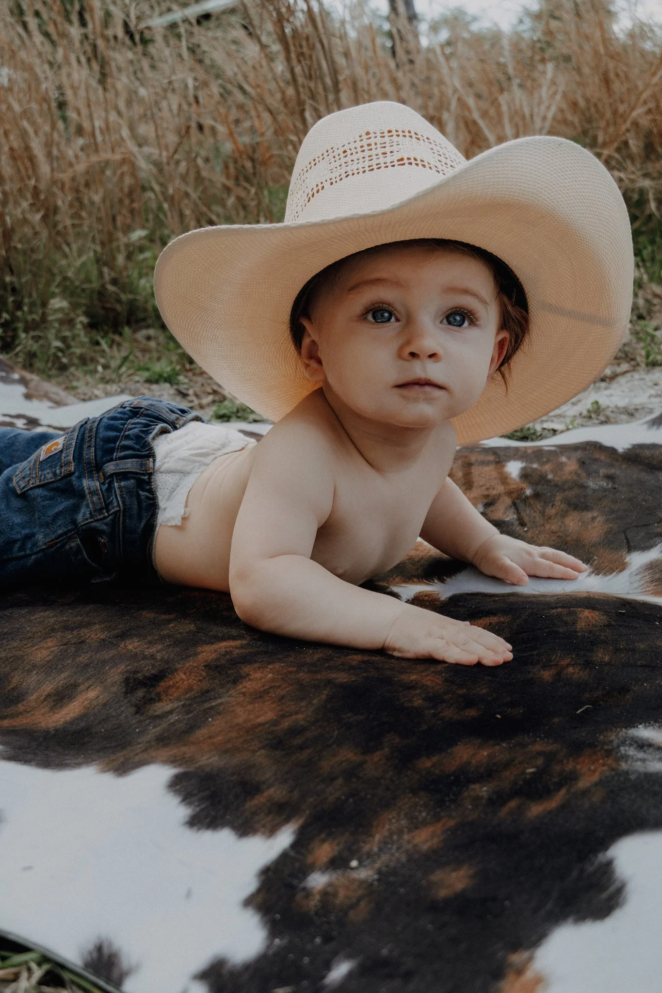 A young child with blue eyes, wearing a large straw sunhat and jeans, lying on their stomach on a cowhide rug outdoors, with a natural background of grasses and trees.
