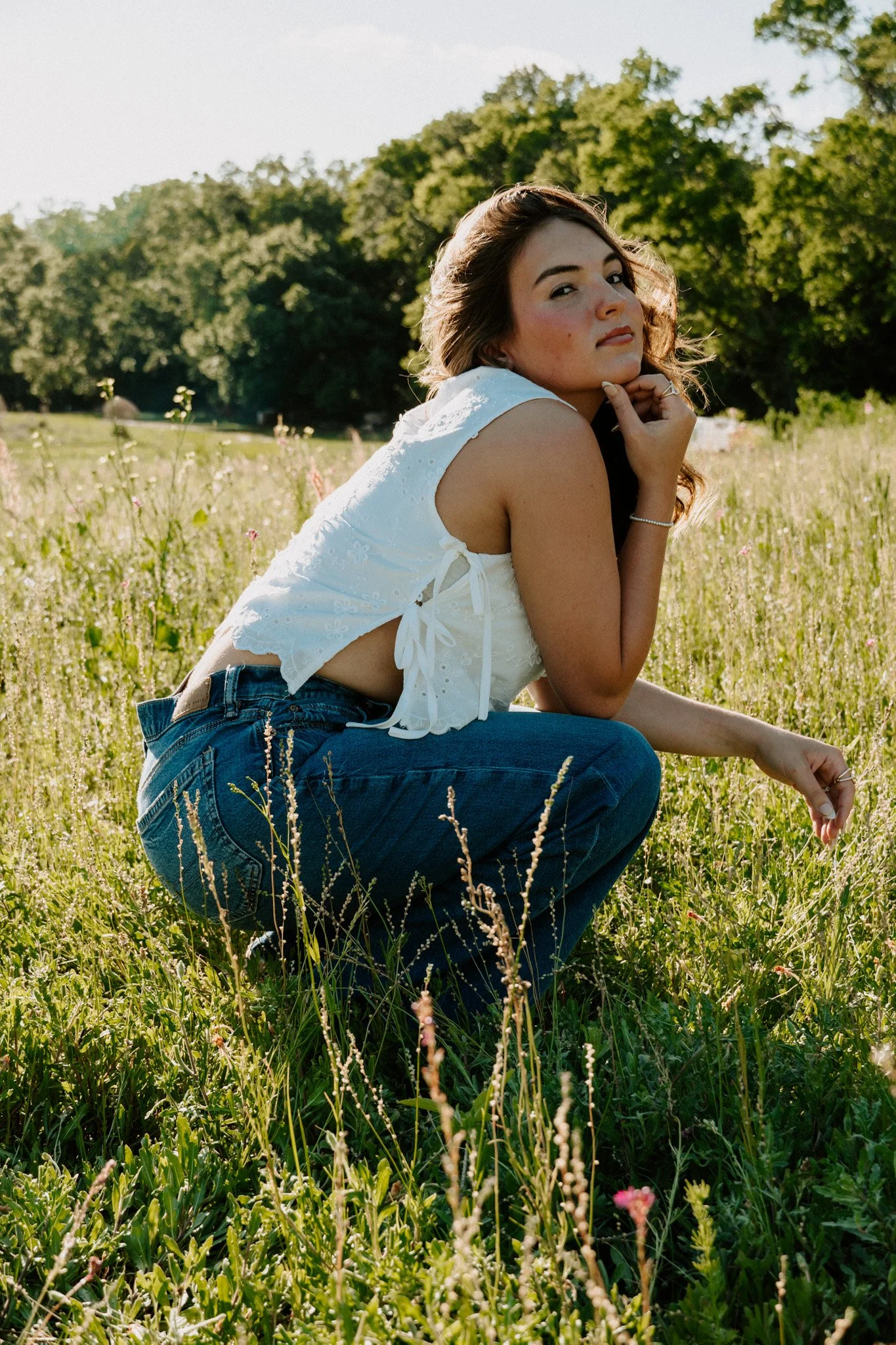 A woman with long brown hair wearing a white sleeveless top and jeans, squatting in a field of tall grass and wildflowers, with trees in the background, during daylight.