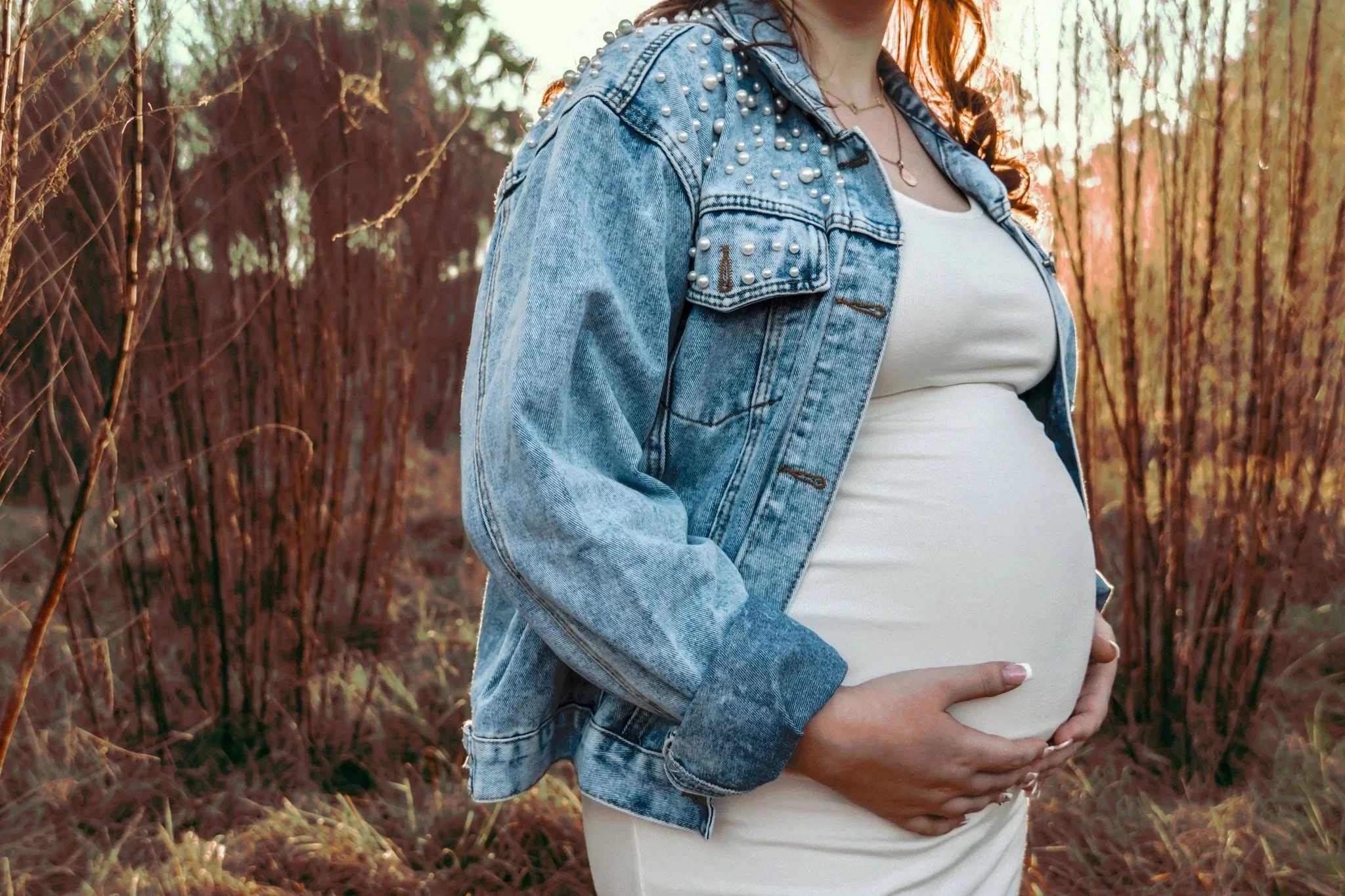 A pregnant woman wearing a white dress and a denim jacket with pearl embellishments, standing outdoors among orange and red grass and tall plants during sunset.