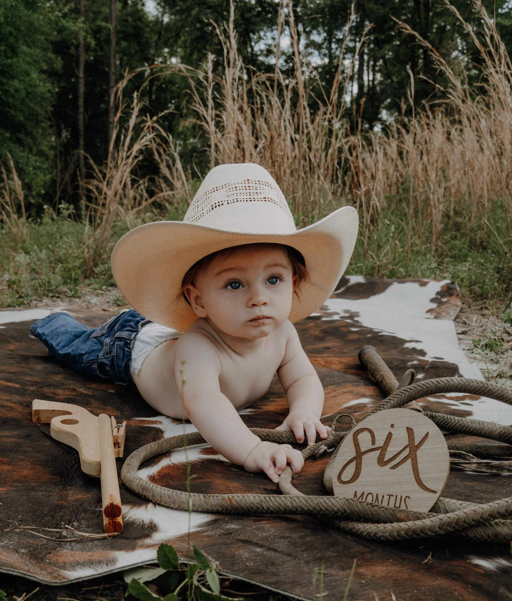 Child lying on their stomach outdoors, wearing a large straw sunhat, surrounded by rustic farm decor, including a wooden cutout with text and thick rope, with tall dried grass and trees in the background.