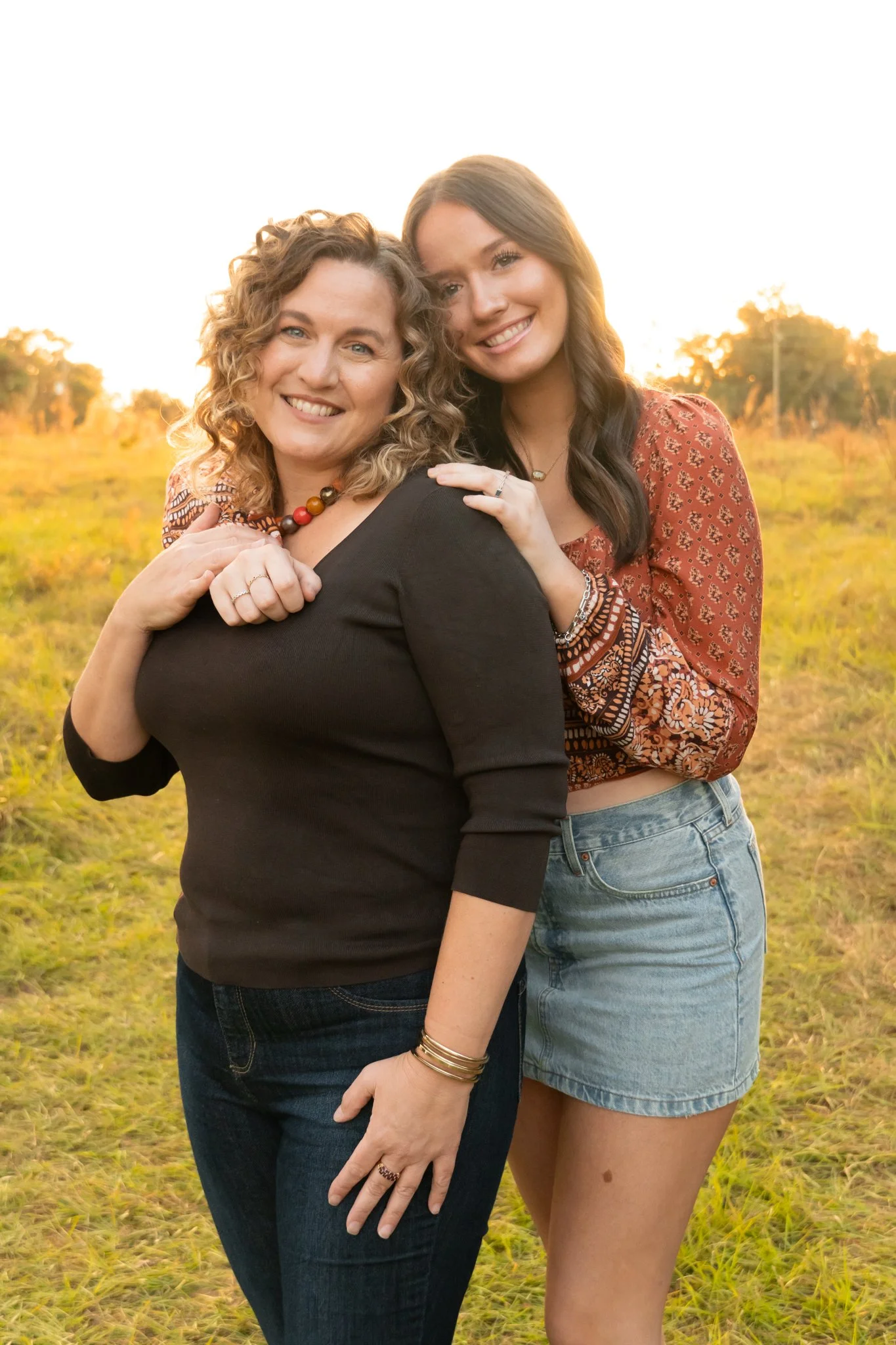 Two women standing outdoors in a grassy field during sunset, smiling and hugging each other. The woman on the left has curly hair, wearing a black top, and the woman on the right has straight hair, wearing a patterned red blouse and denim shorts.