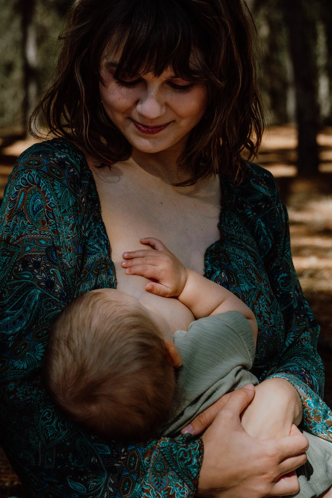 A woman with shoulder-length dark brown hair and a patterned dress is breastfeeding a young child in a forest setting.