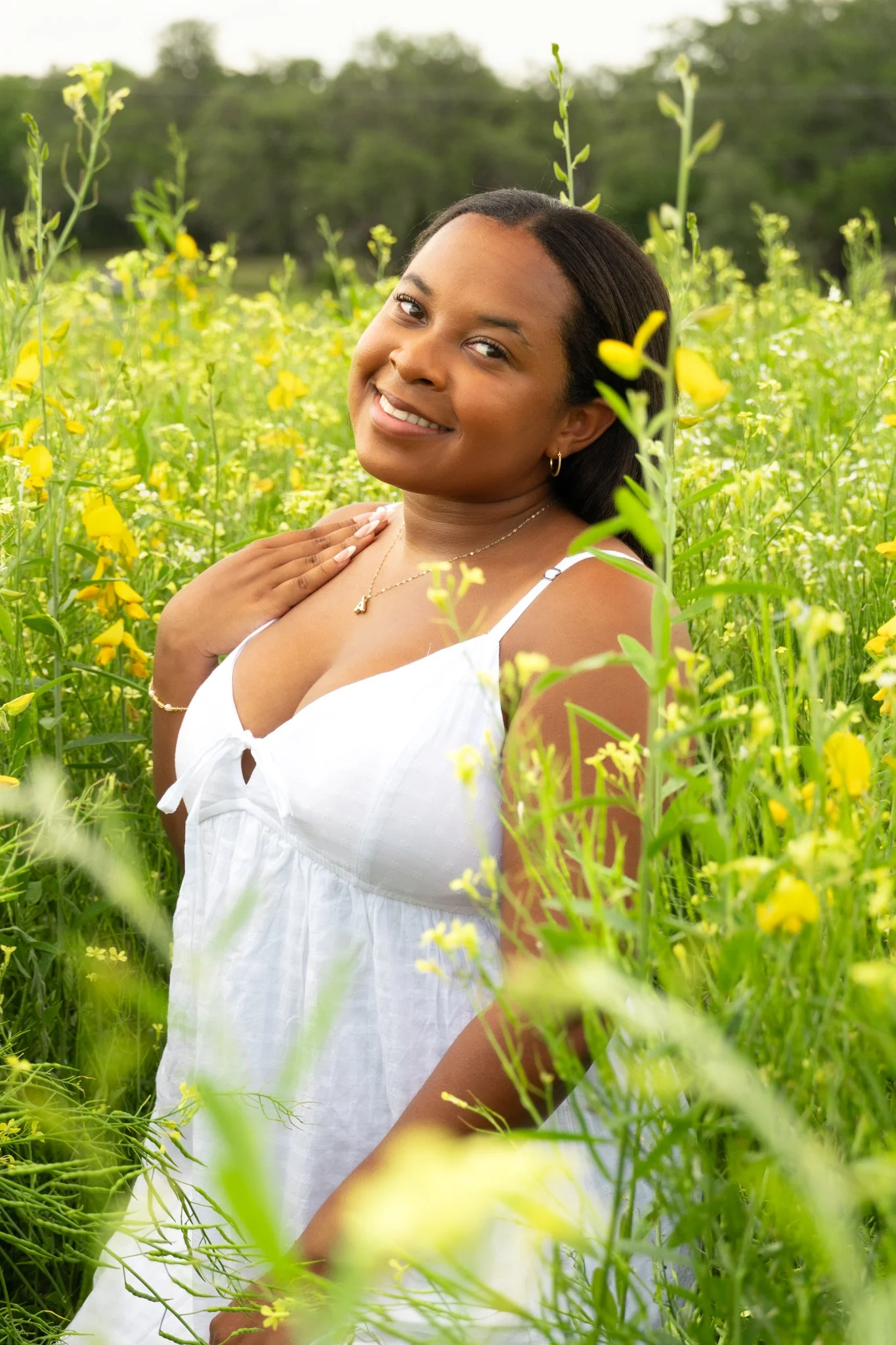 A woman in a white dress standing amidst yellow flowers in a lush green field, smiling softly at the camera.