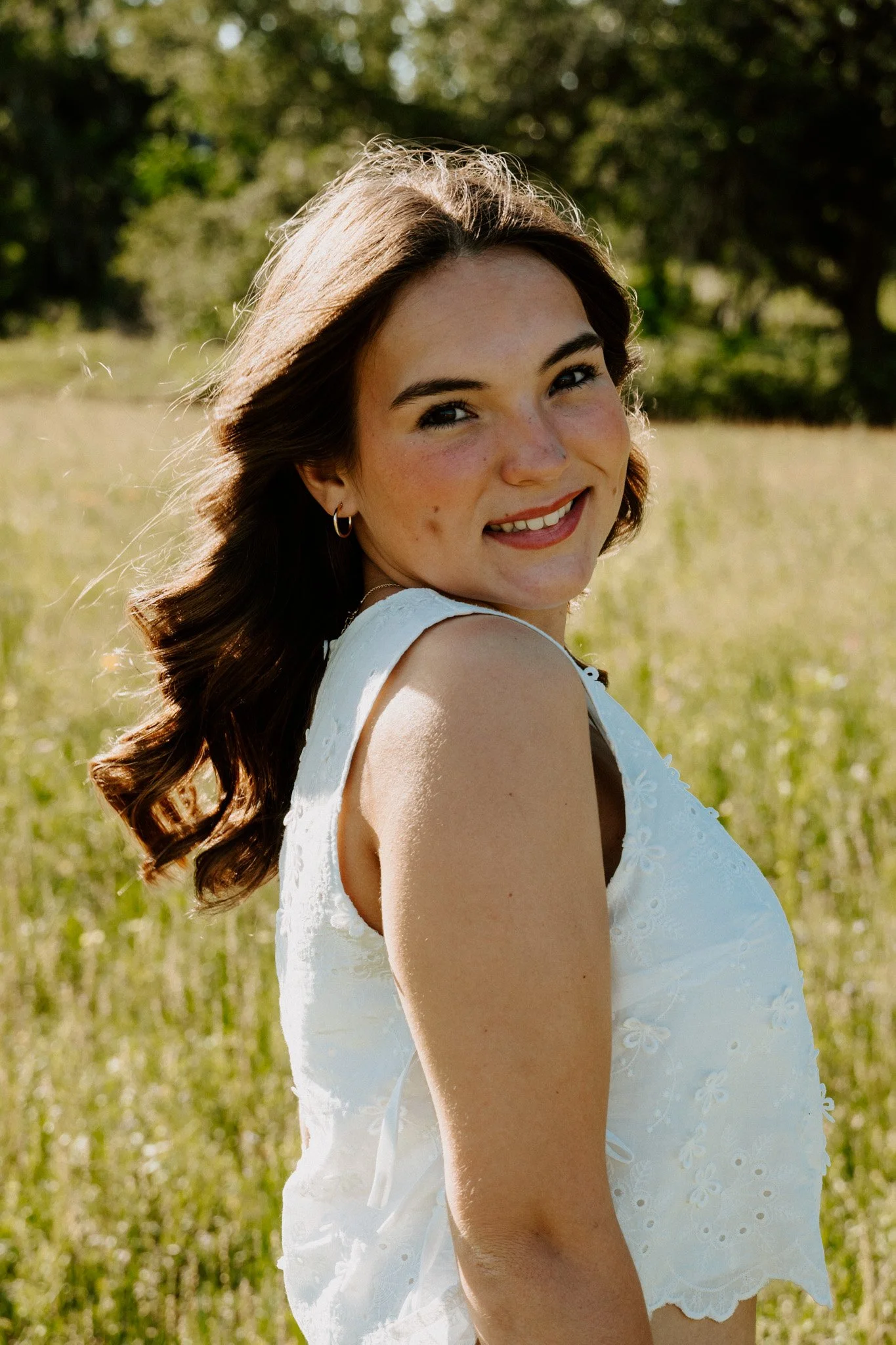 A young woman with long, wavy brown hair and a bright smile outdoors in a sunlit grassy field, wearing a white sleeveless dress with floral embroidery.