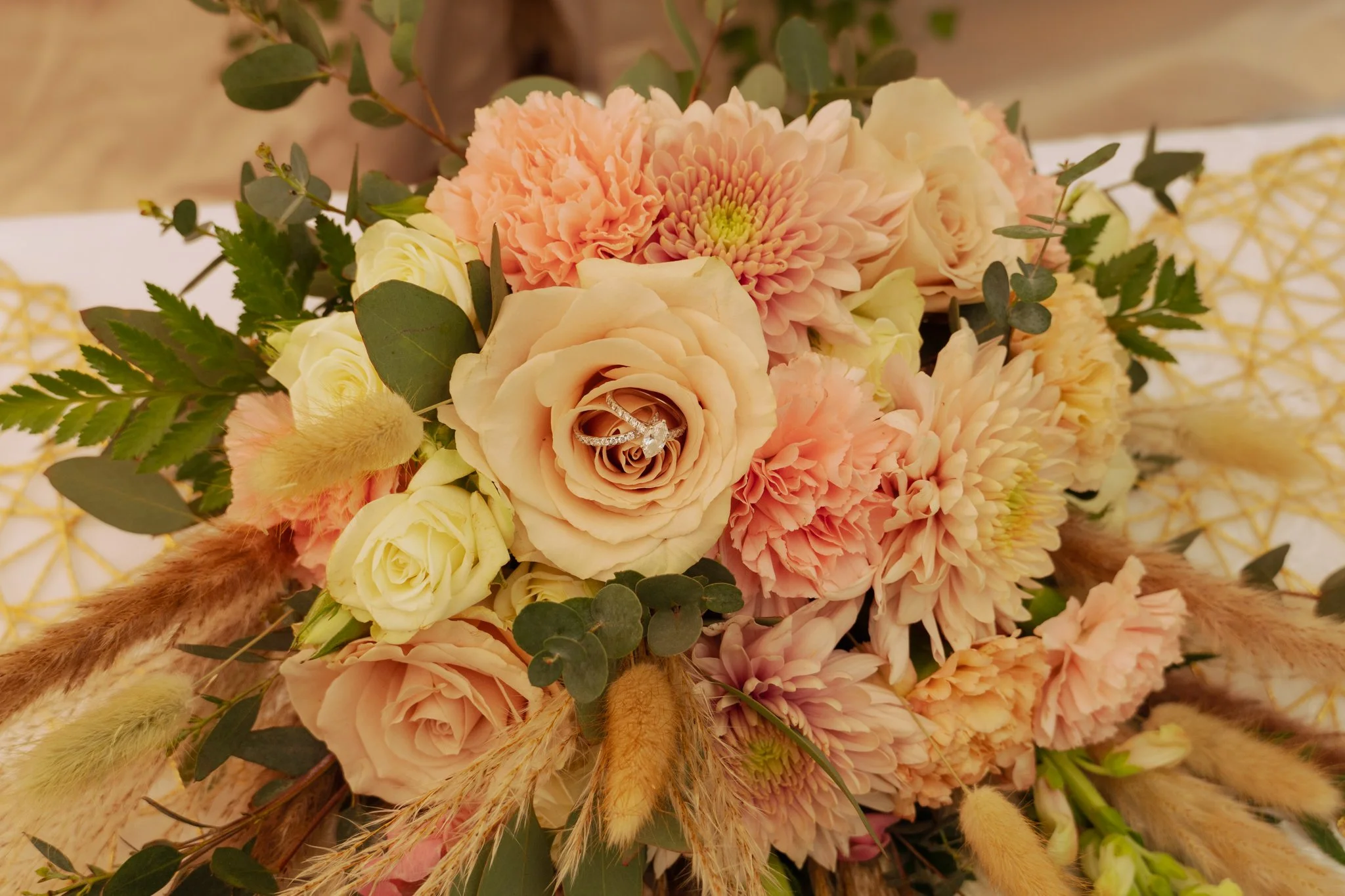 A floral arrangement with peach and cream-colored roses, pink carnations, greenery, and dried grass, with a diamond engagement ring placed inside a cream rose.