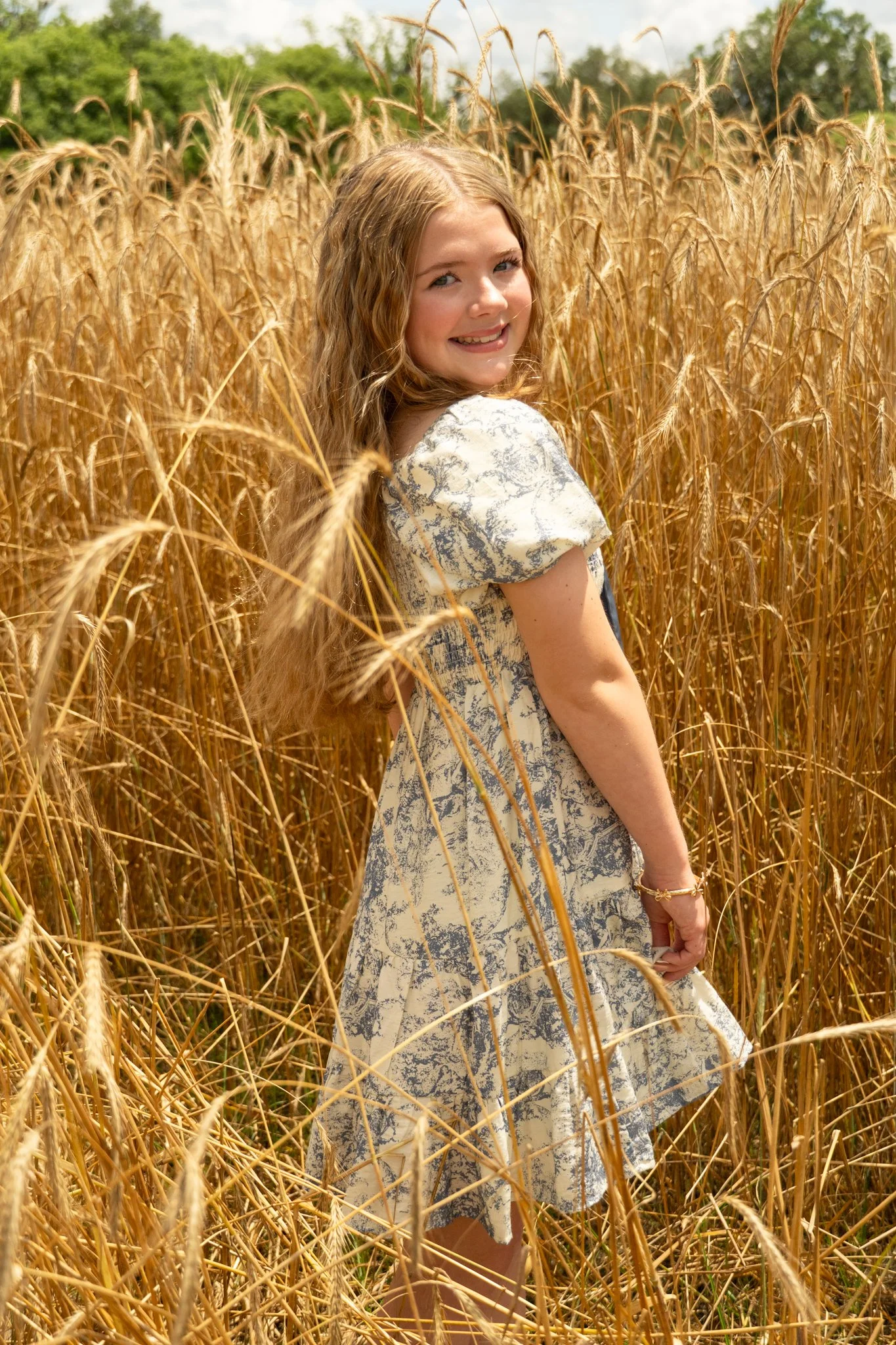 A young woman with long, wavy blonde hair smiling and looking over her shoulder while standing in a golden wheat field on a sunny day.
