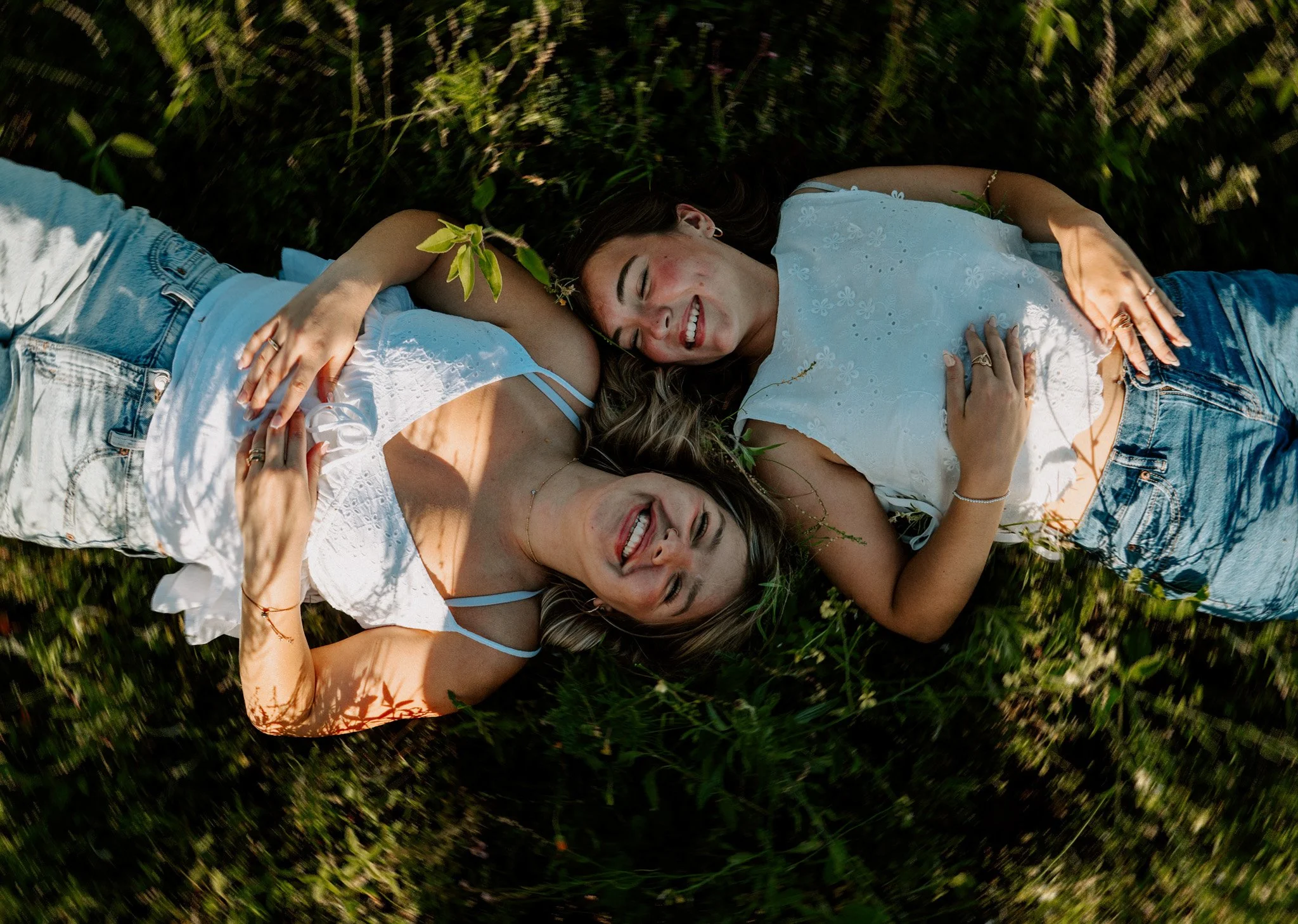Three women lying on grass, smiling, with one woman resting her head on another's shoulder, enjoying a sunny day.