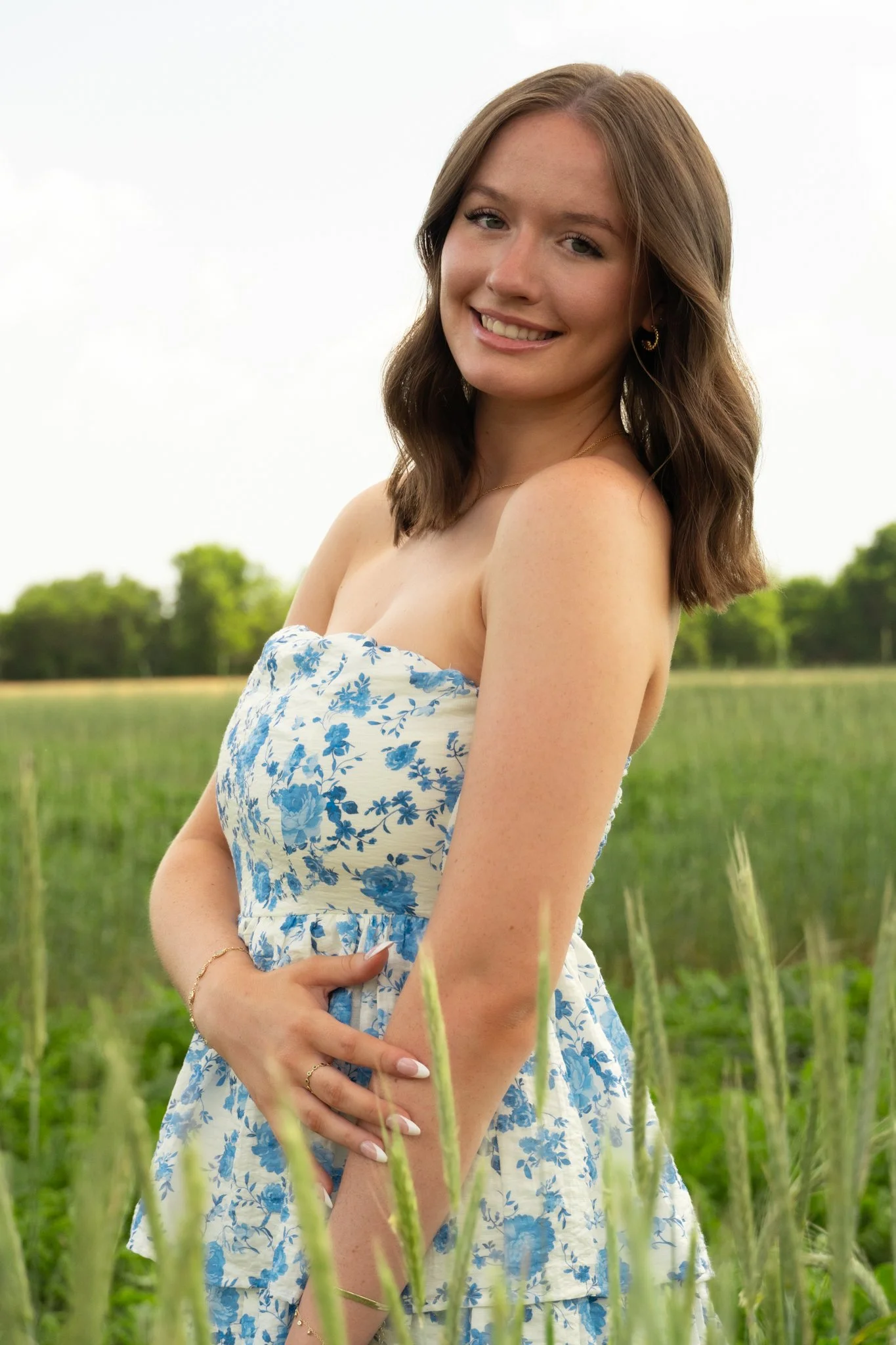 A young woman with shoulder-length brown hair, smiling, standing outdoors in a green field with trees in the background, wearing a blue and white floral dress.