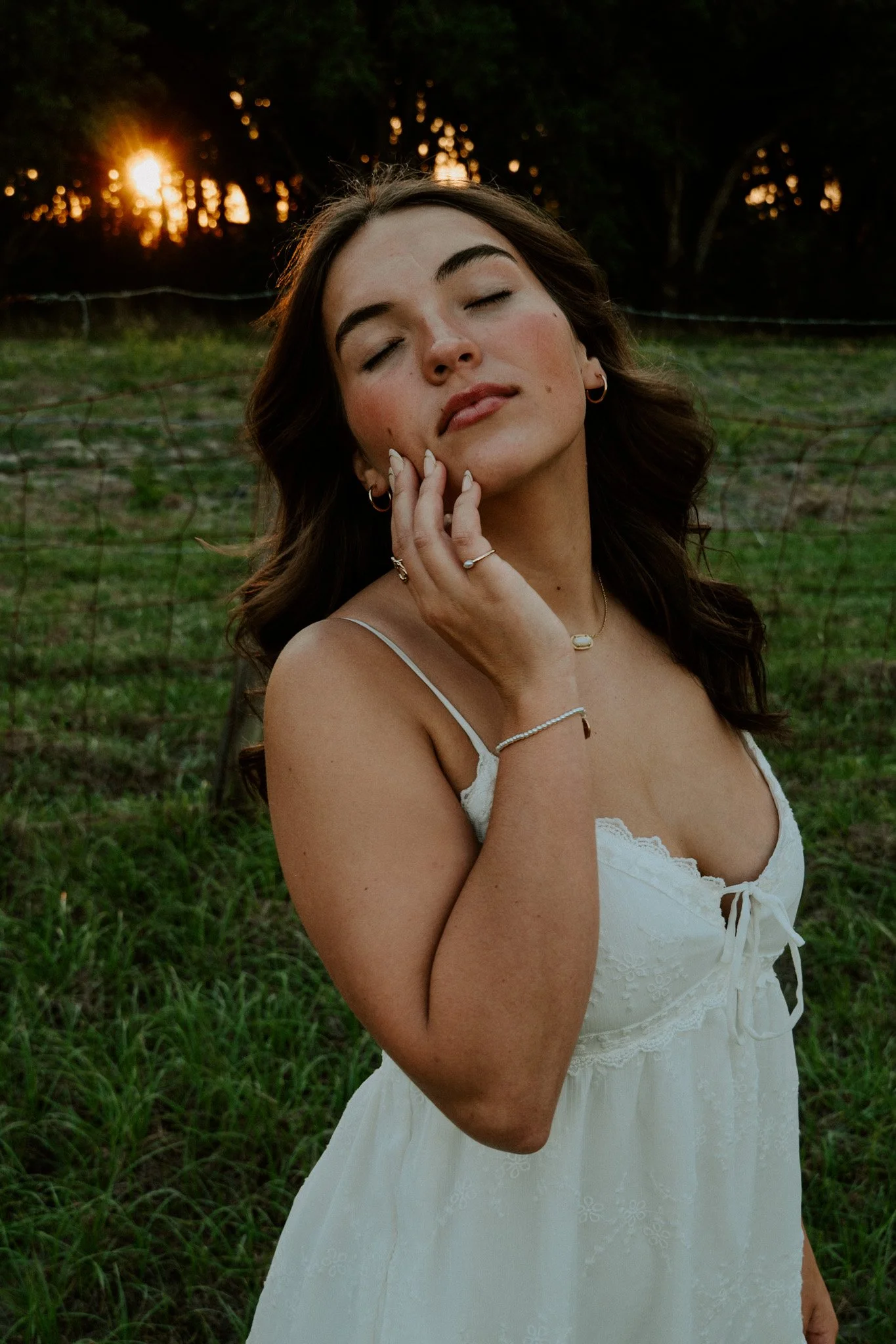 A woman with long brown hair and hoop earrings gently touching her face, standing outdoors at sunset with trees and grass in the background.