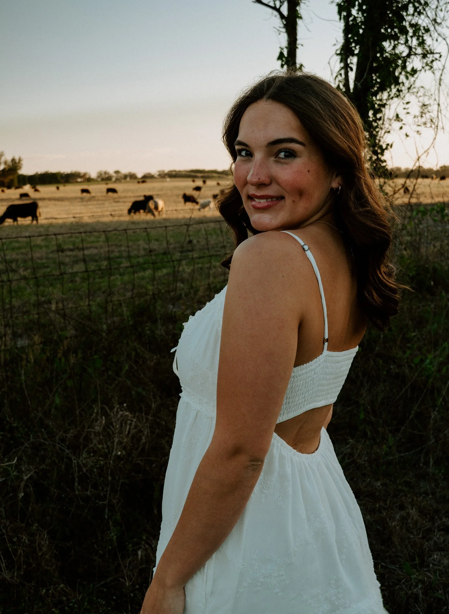 A woman with brown hair and earrings wearing a white dress, standing outdoors near a fence with cows grazing in the background during sunset.