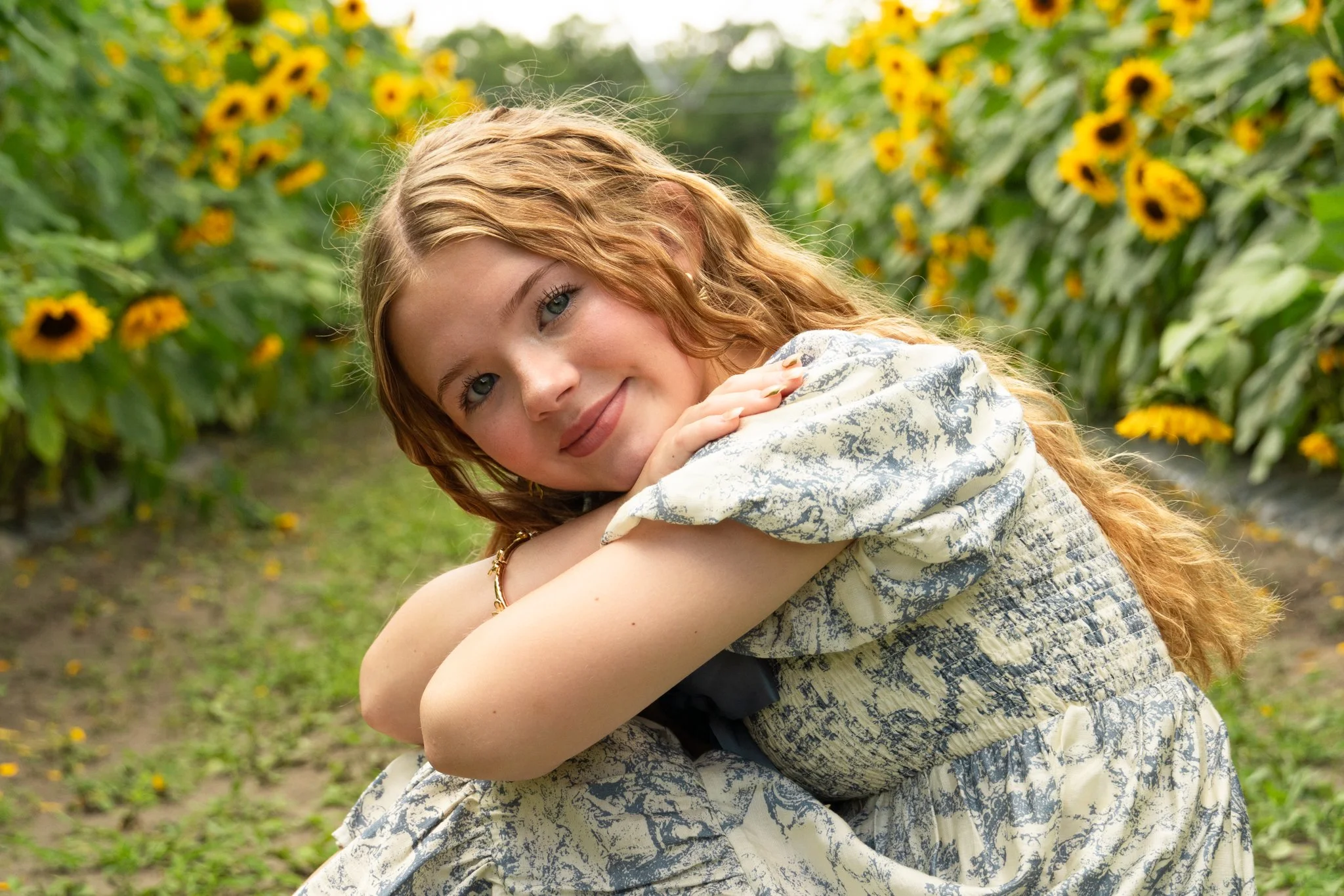 A woman with red hair and blue eyes sitting in a sunflower field, smiling and pretending to sleep.