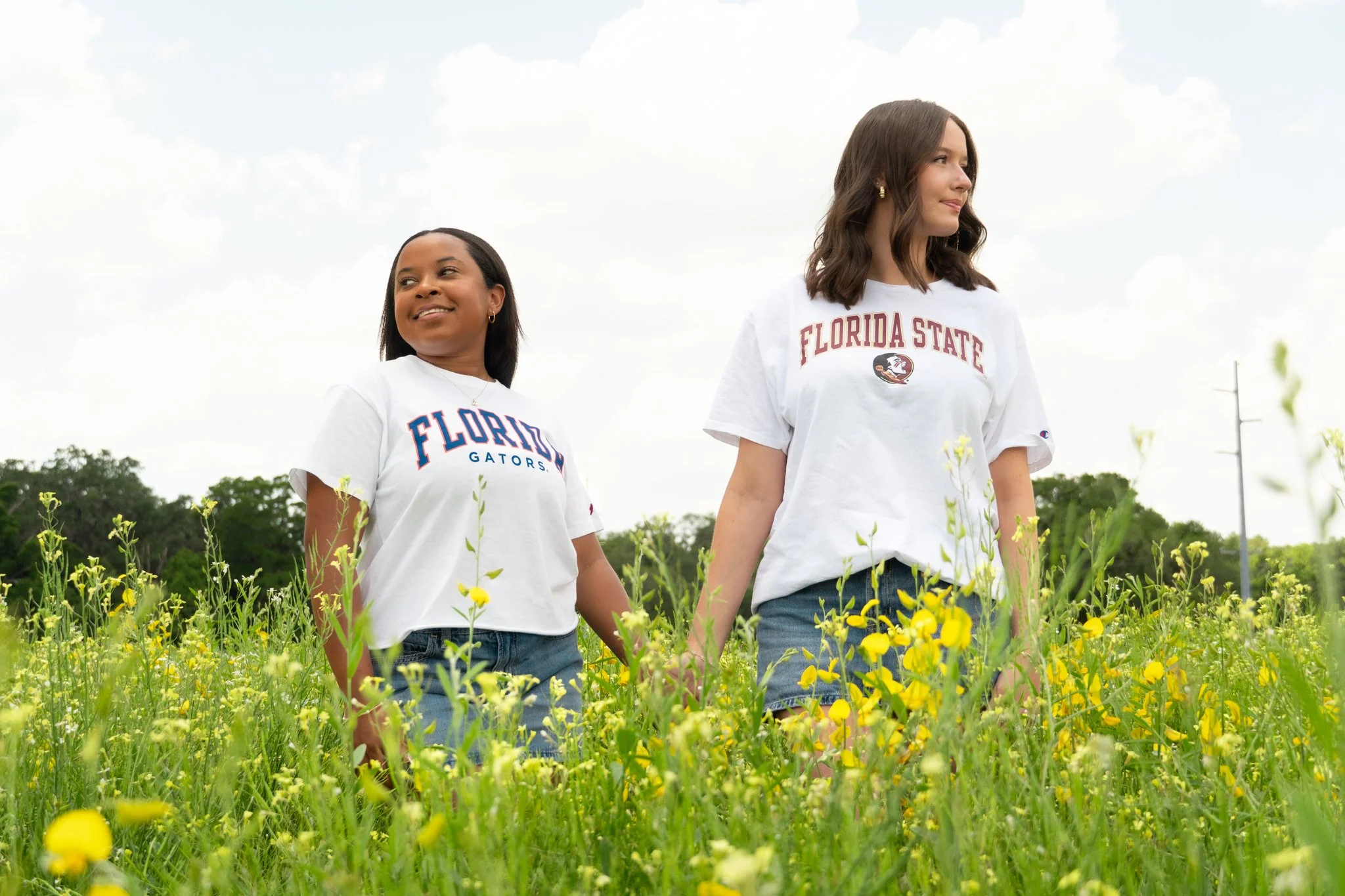 Two women walking hand in hand through a field of yellow flowers, wearing white T-shirts with Florida State University logos.
