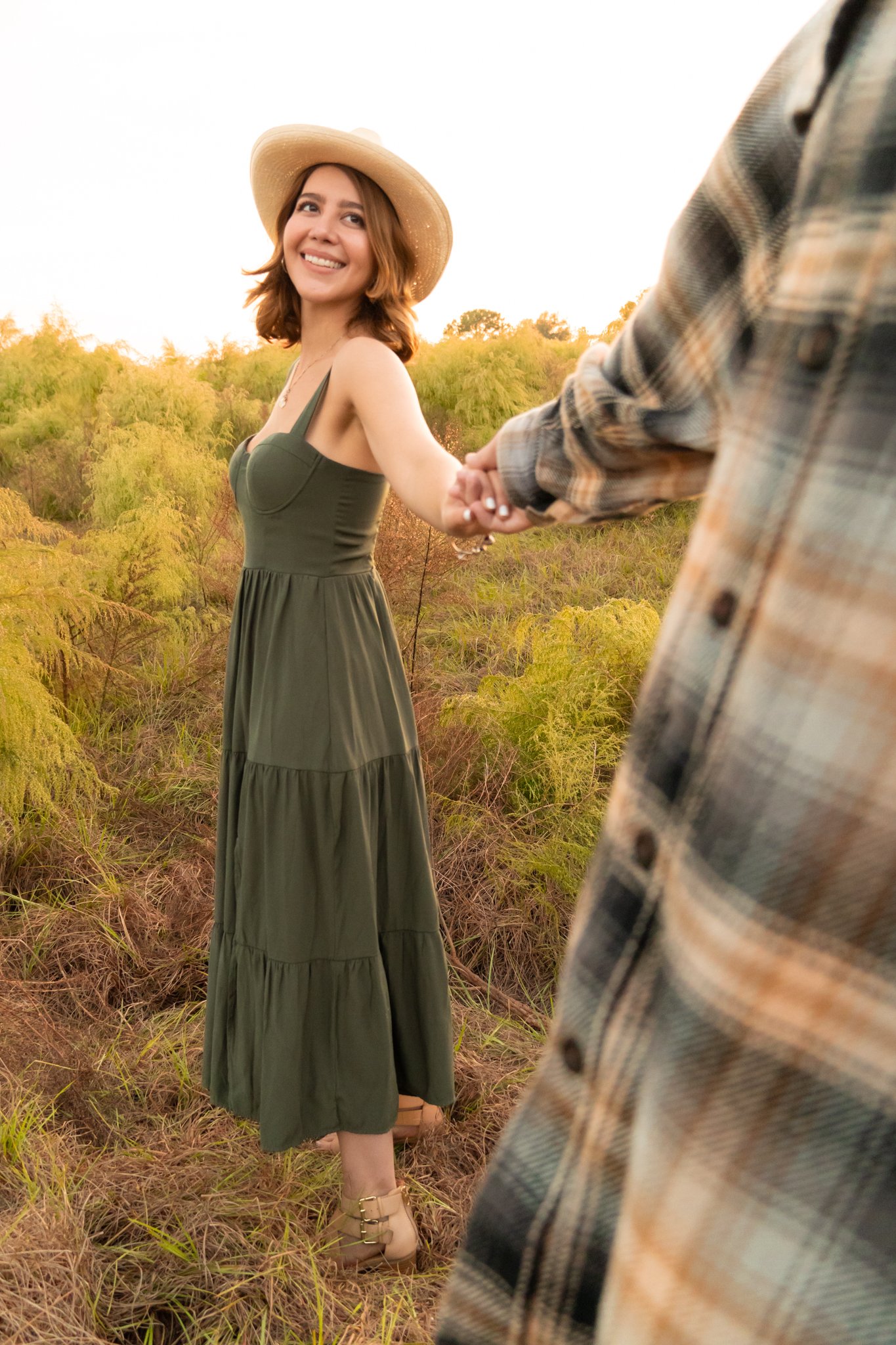 A woman in a green dress and a wide-brimmed straw hat is holding hands with a person wearing a plaid shirt in a natural outdoor setting with green trees and grass.